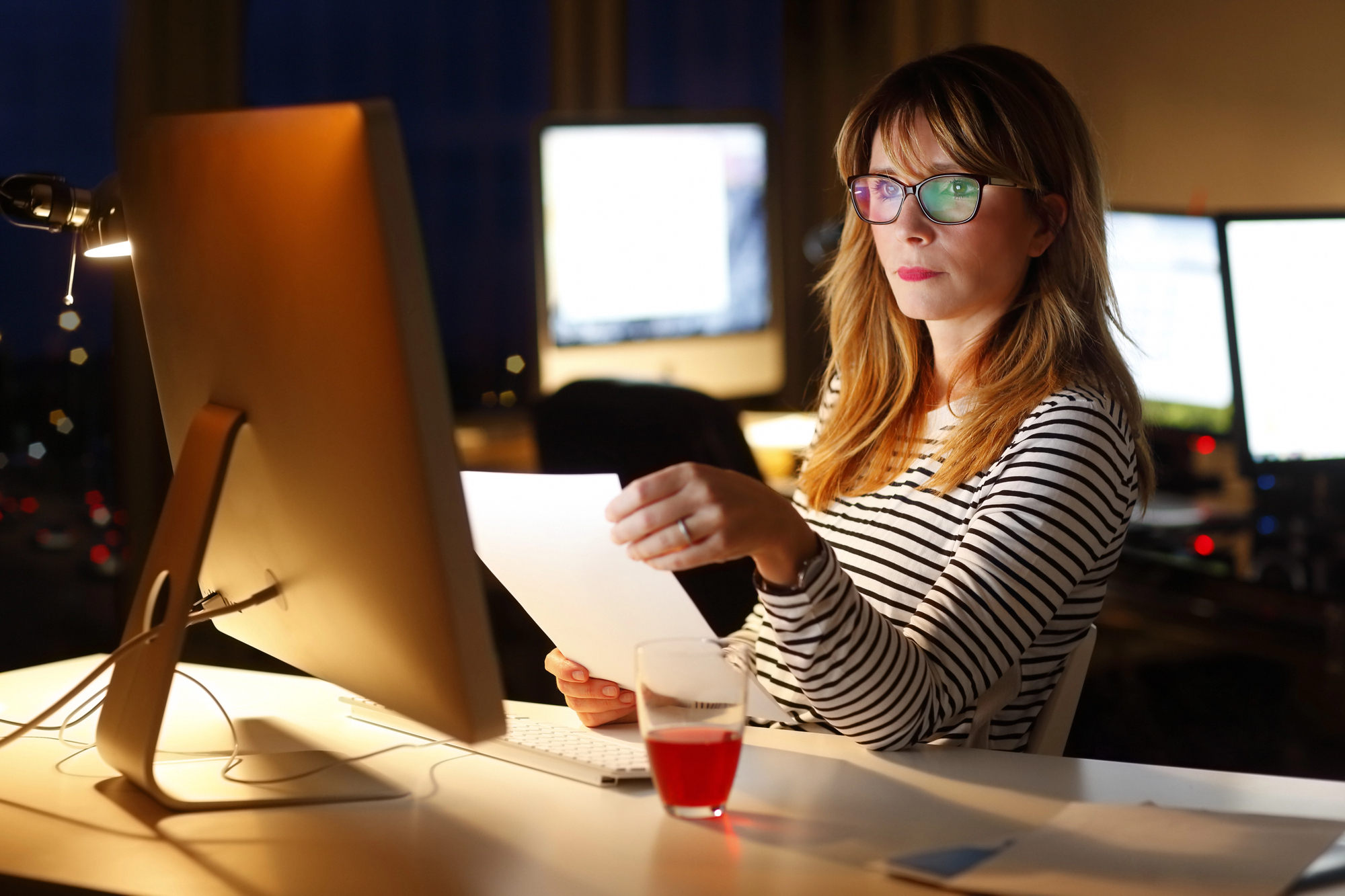 A woman wearing glasses and a striped shirt sits at a desk, focused on a computer screen. She holds a sheet of paper and has a glass with a red drink nearby. The background features illuminated screens and a dimly lit room.