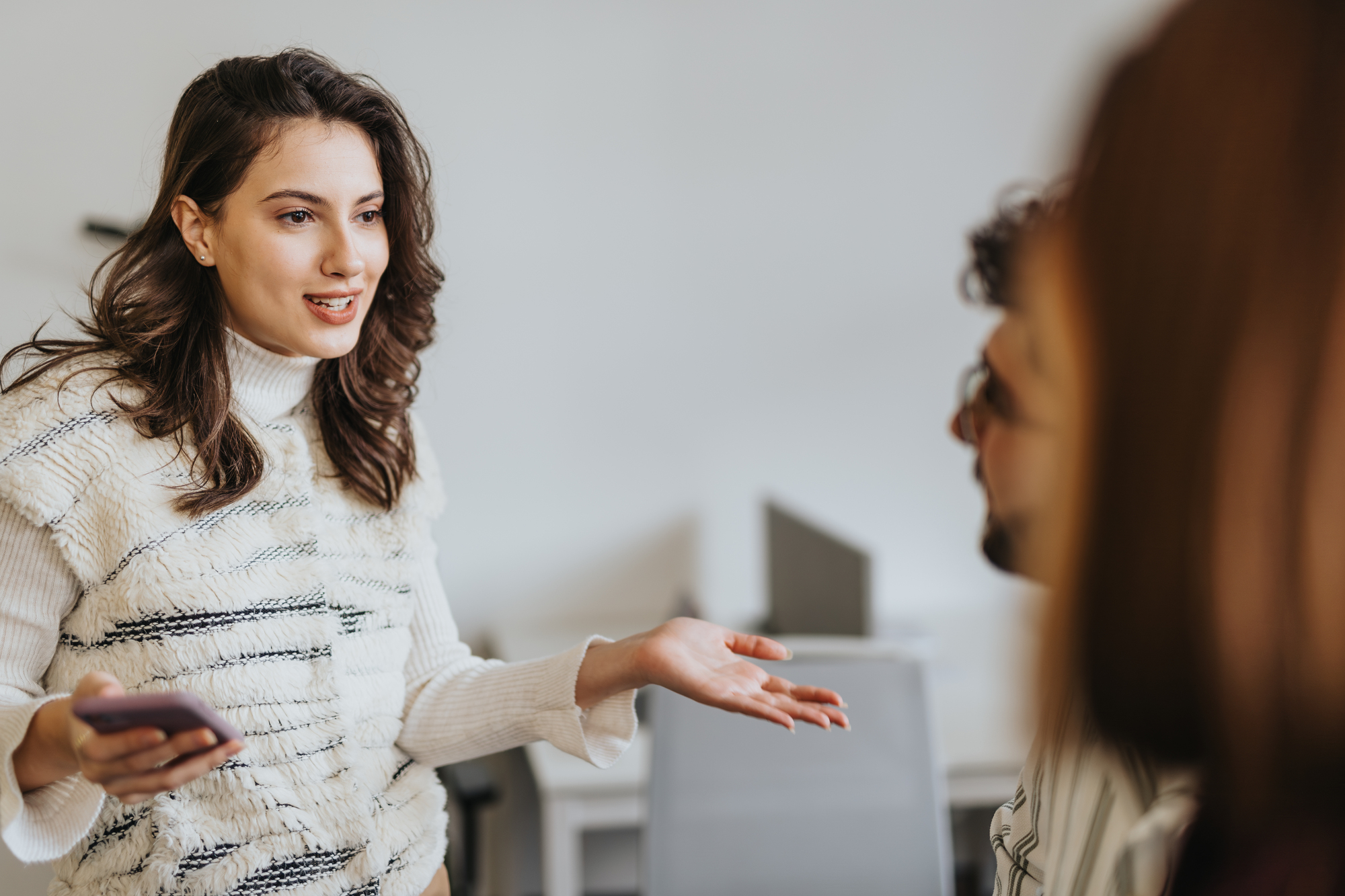 A woman in a white turtleneck sweater gestures while holding a smartphone, engaging in conversation with two people whose backs are partly visible. They are in a bright office setting with desks and chairs in the background.