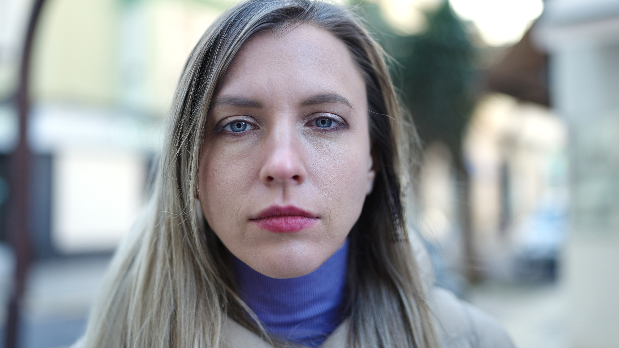 A woman with long blonde hair and a serious expression stands outdoors. She is wearing a purple turtleneck and a beige jacket. The background is blurred, with hints of buildings and trees.