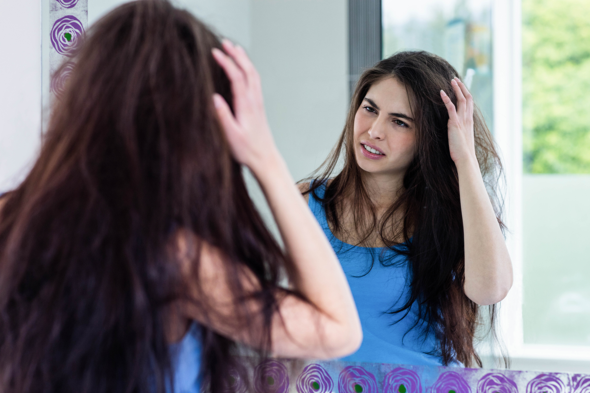 A woman with long, dark hair stands in front of a mirror, wearing a blue tank top. She looks at her reflection with a slightly confused expression while adjusting her hair. The background features a window with greenery visible outside.