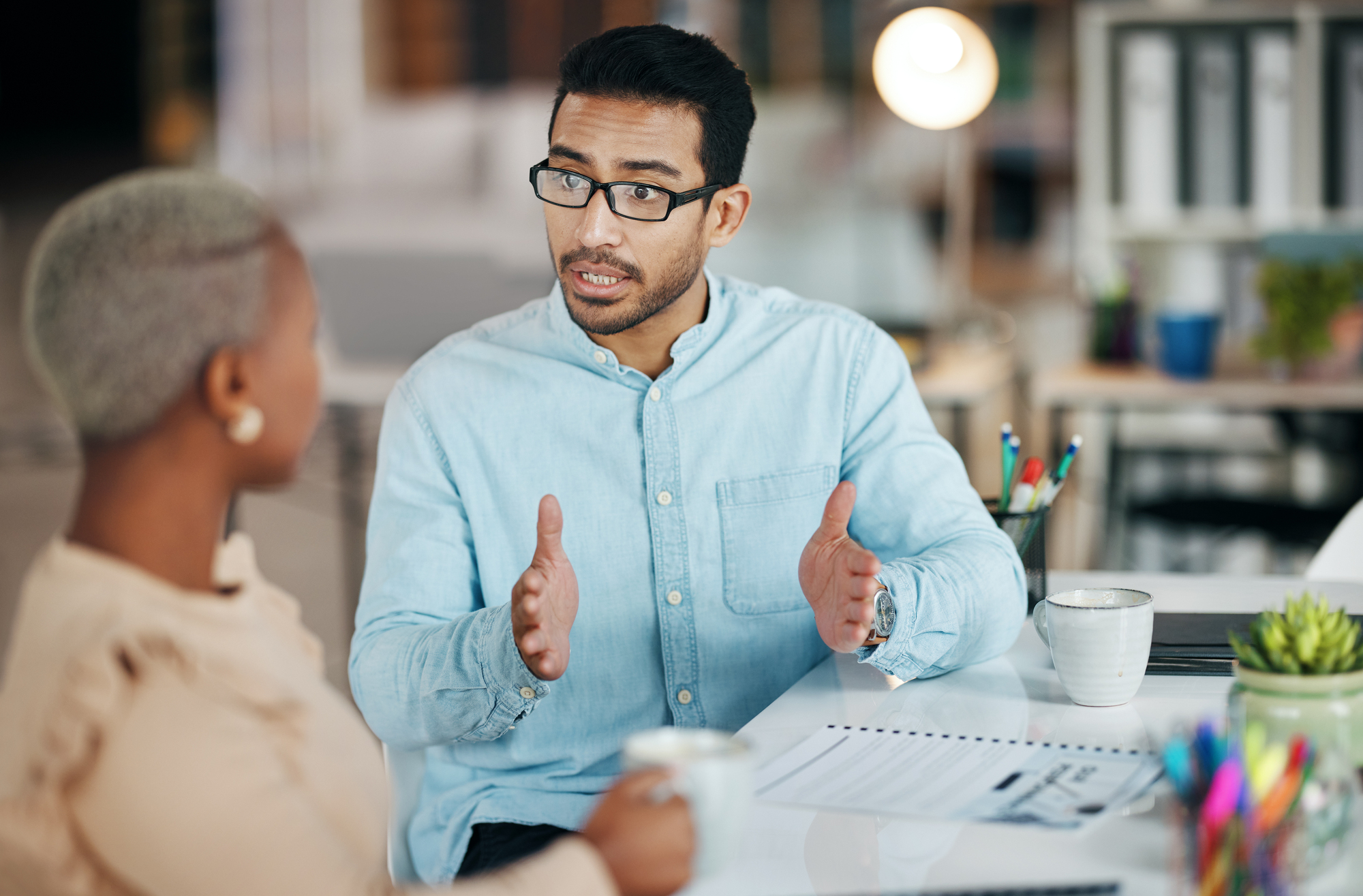 A man in glasses and a blue shirt is gesturing while talking to a woman with short hair in an office setting. They are seated at a table with coffee mugs, documents, and colorful pens in the foreground.