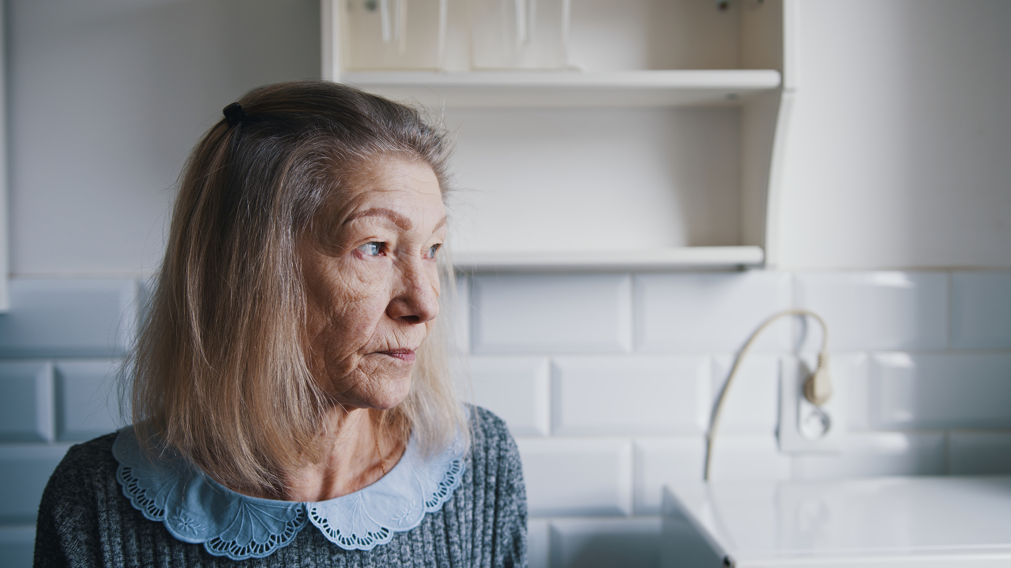 An elderly woman with long hair looks contemplatively to the side while sitting in a bright kitchen with white tiled walls and shelves.