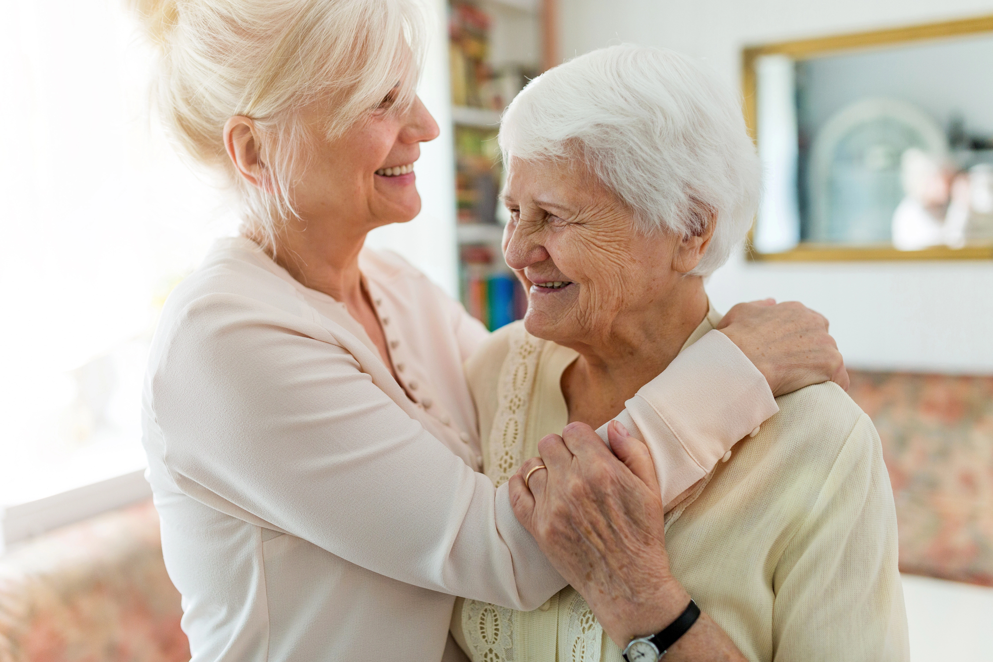 Two elderly women are embracing and smiling warmly at each other in a cozy room. Sunlight brightens the scene, highlighting their joyful expressions. One woman has blonde hair, and the other has white hair. A mirror and shelves are in the background.