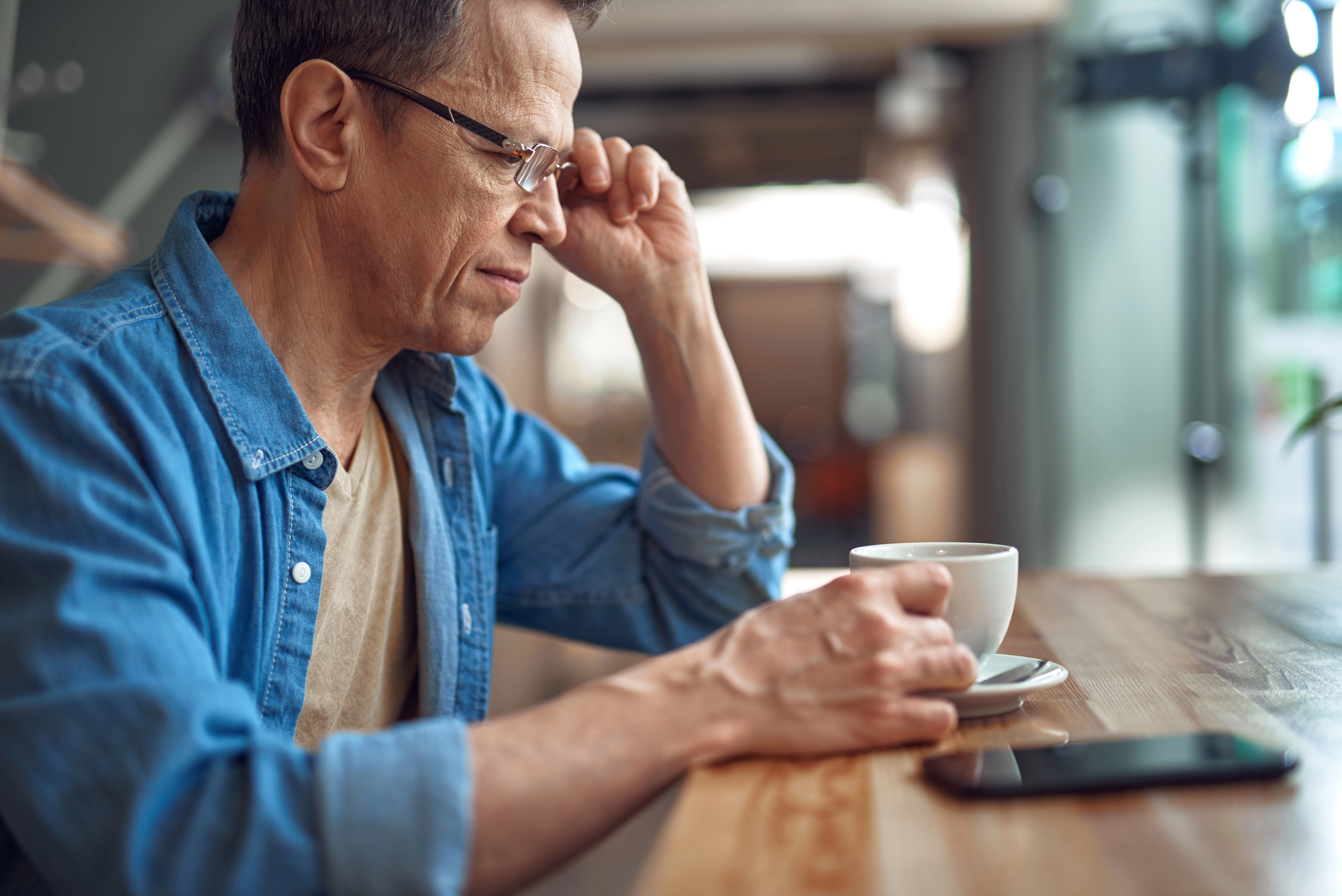 A person wearing glasses and a denim shirt sits at a wooden table in a cozy café, holding a cup of coffee. A smartphone rests on the table nearby. The background is softly blurred, creating a warm and inviting atmosphere.