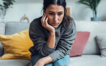 A woman sits on a sofa, leaning forward with her chin resting on her hand. She appears thoughtful or concerned. The room has colorful cushions and a blurred plant in the background, creating a cozy atmosphere.