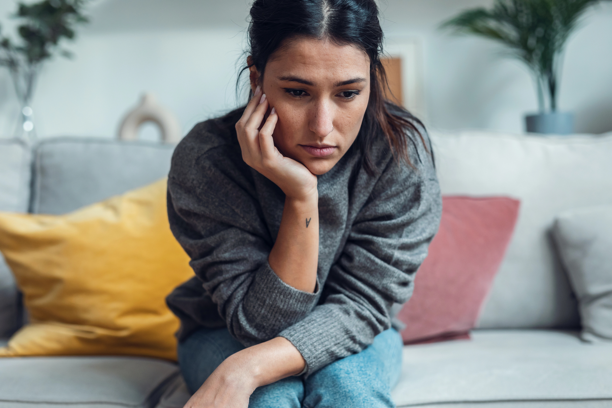 A woman sits on a sofa, leaning forward with her chin resting on her hand. She appears thoughtful or concerned. The room has colorful cushions and a blurred plant in the background, creating a cozy atmosphere.