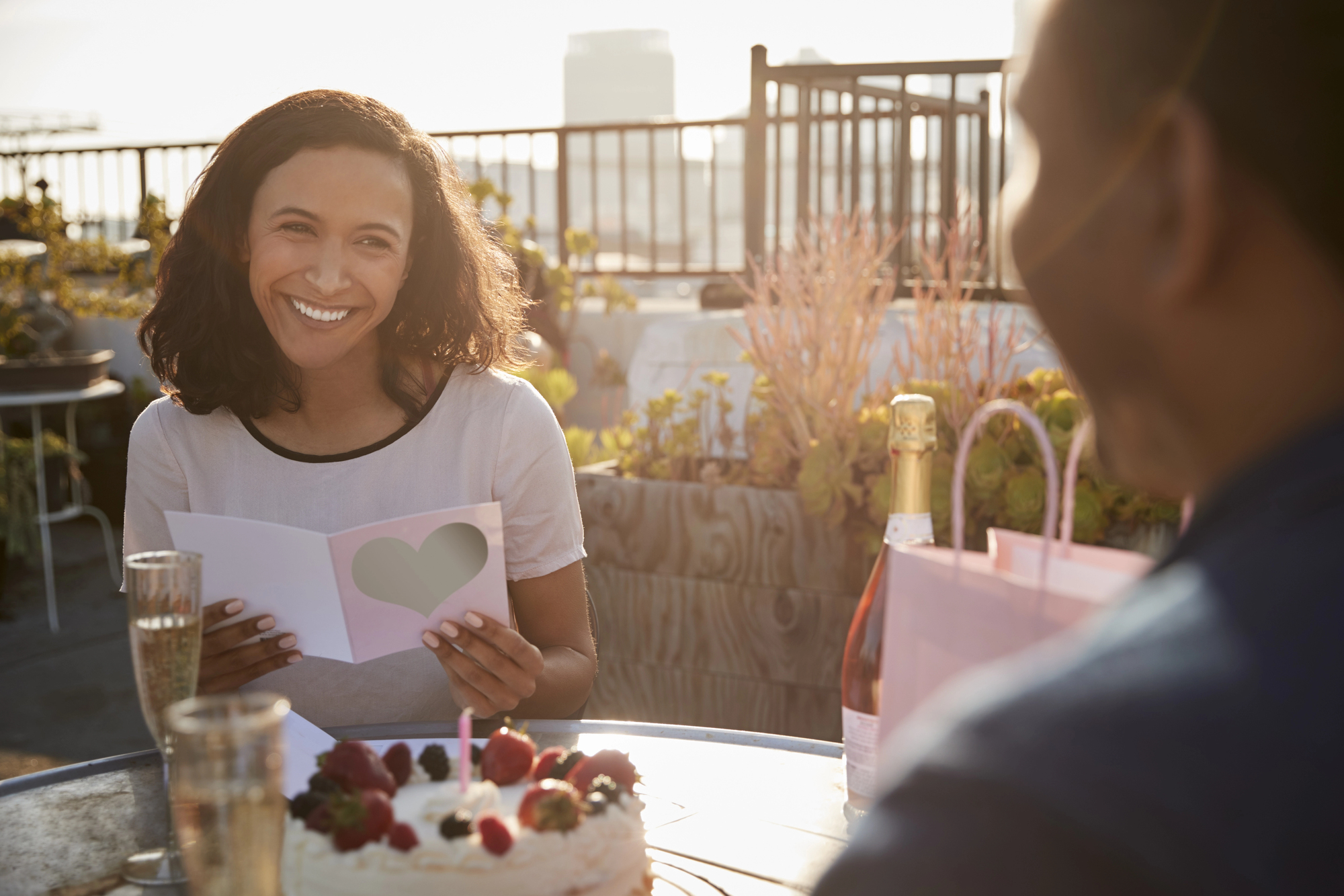 A woman smiles while holding a card with a heart on it. She is seated at a table with a birthday cake topped with fruit, two glasses of champagne, and a gift bag. It's a sunny day on a rooftop patio with plants in the background.