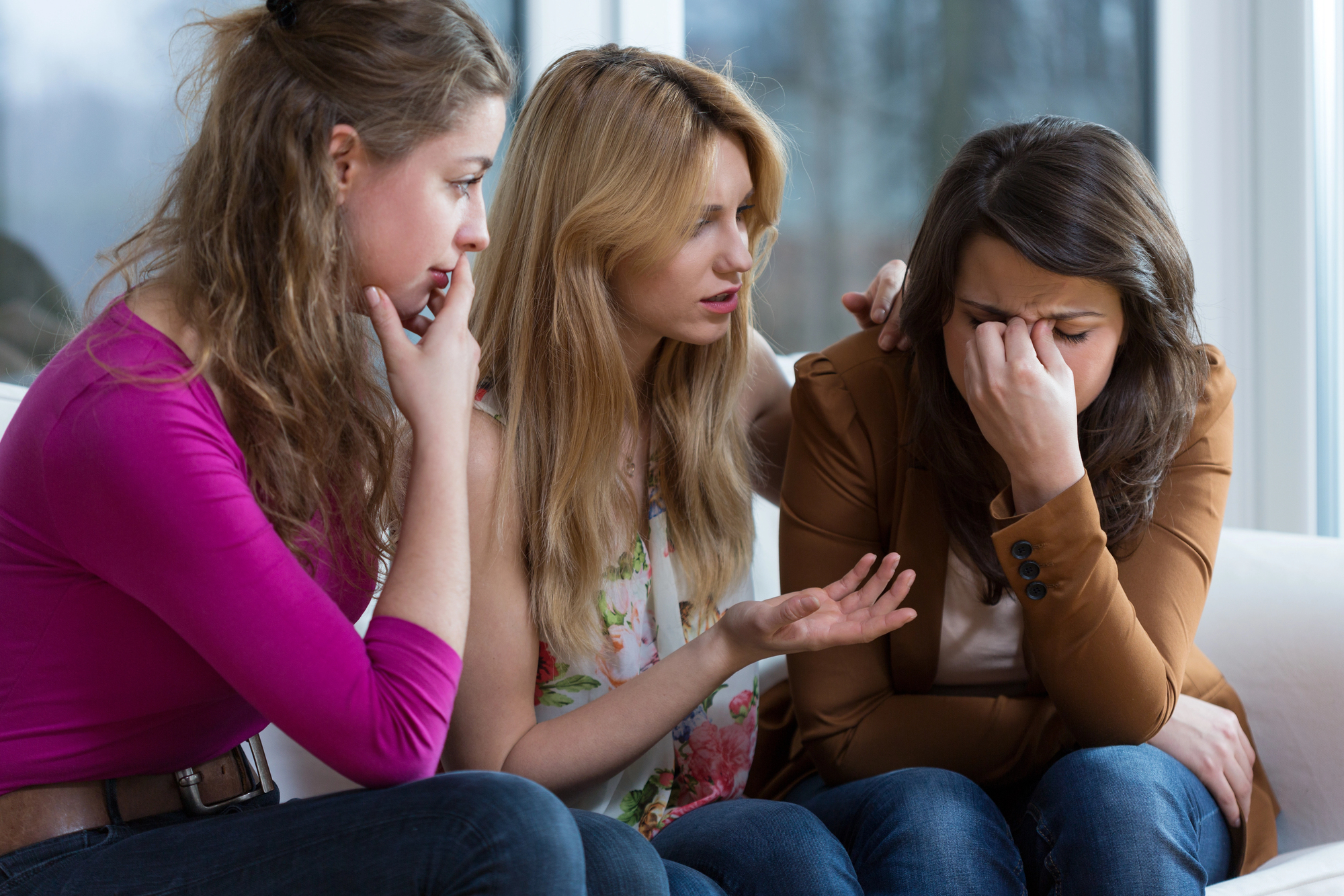 Three women are sitting together on a couch. The woman on the right appears upset, covering her face with one hand. The other two women are offering comfort and support, with one speaking and the other placing a hand on her shoulder.