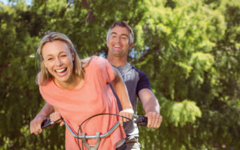 A smiling woman wearing a pink shirt is riding a bicycle with a man in a grey shirt behind her in a park. Both are laughing and enjoying the sunny day, surrounded by green trees.