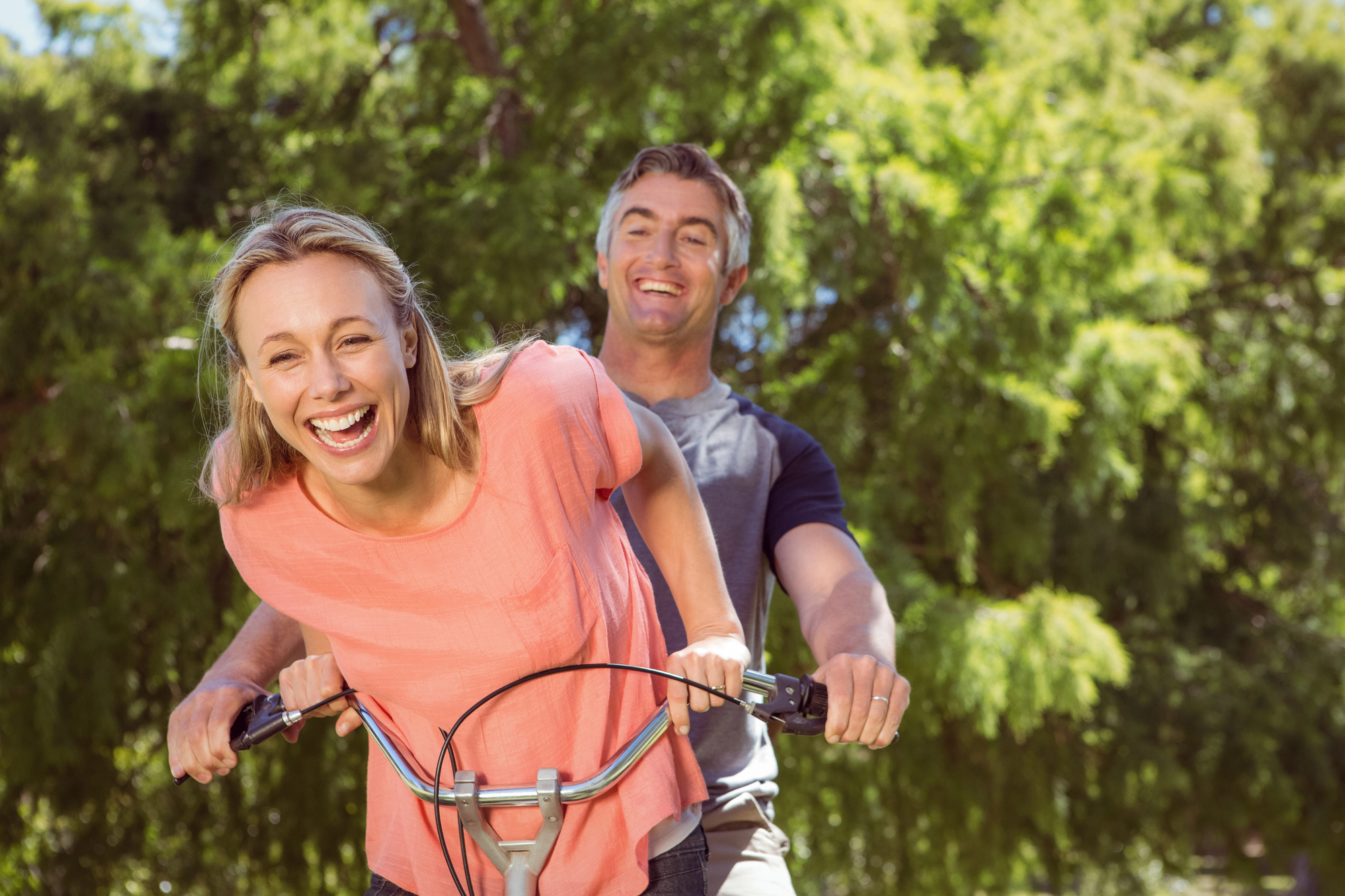 A smiling woman wearing a pink shirt is riding a bicycle with a man in a grey shirt behind her in a park. Both are laughing and enjoying the sunny day, surrounded by green trees.