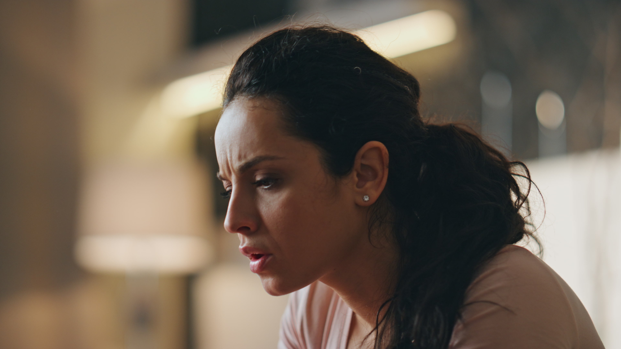 A woman with long, dark hair in a ponytail looks focused and concerned. She is wearing a light pink top and earrings. The background is softly blurred, indicating an indoor setting.