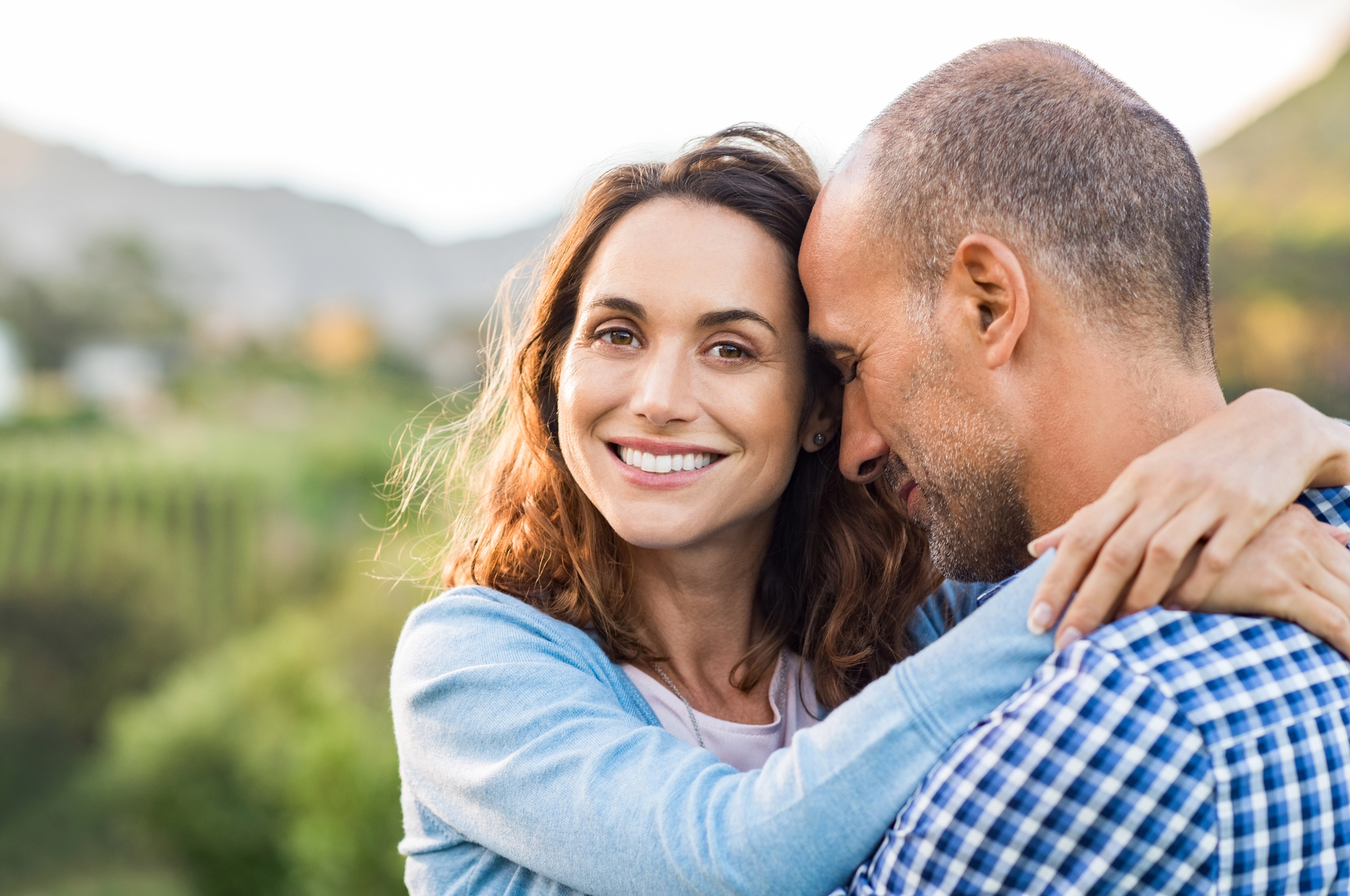 A woman smiling and embracing a man with her arms around his neck. They are standing outdoors, with greenery and mountainous landscape in the background. The man is wearing a checkered shirt, and the woman is in a light-colored top and cardigan.