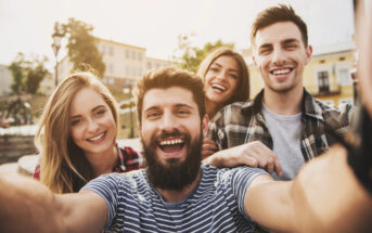 Four smiling friends gather closely for a selfie outdoors in a sunny urban setting. The group, consisting of two men and two women, appears happy and joyful, wearing casual clothes and standing in front of buildings with a clear sky in the background.