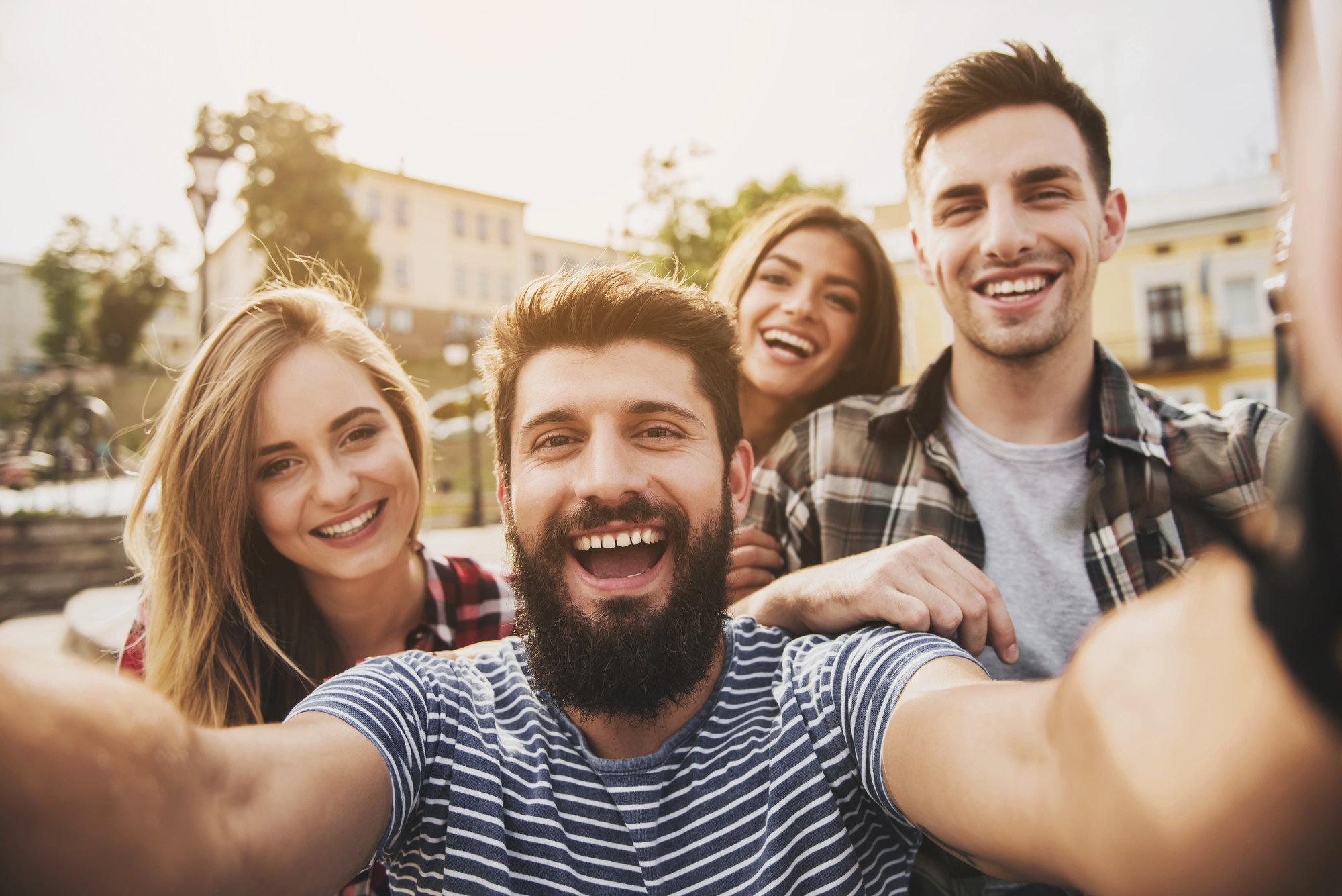 Four smiling friends gather closely for a selfie outdoors in a sunny urban setting. The group, consisting of two men and two women, appears happy and joyful, wearing casual clothes and standing in front of buildings with a clear sky in the background.