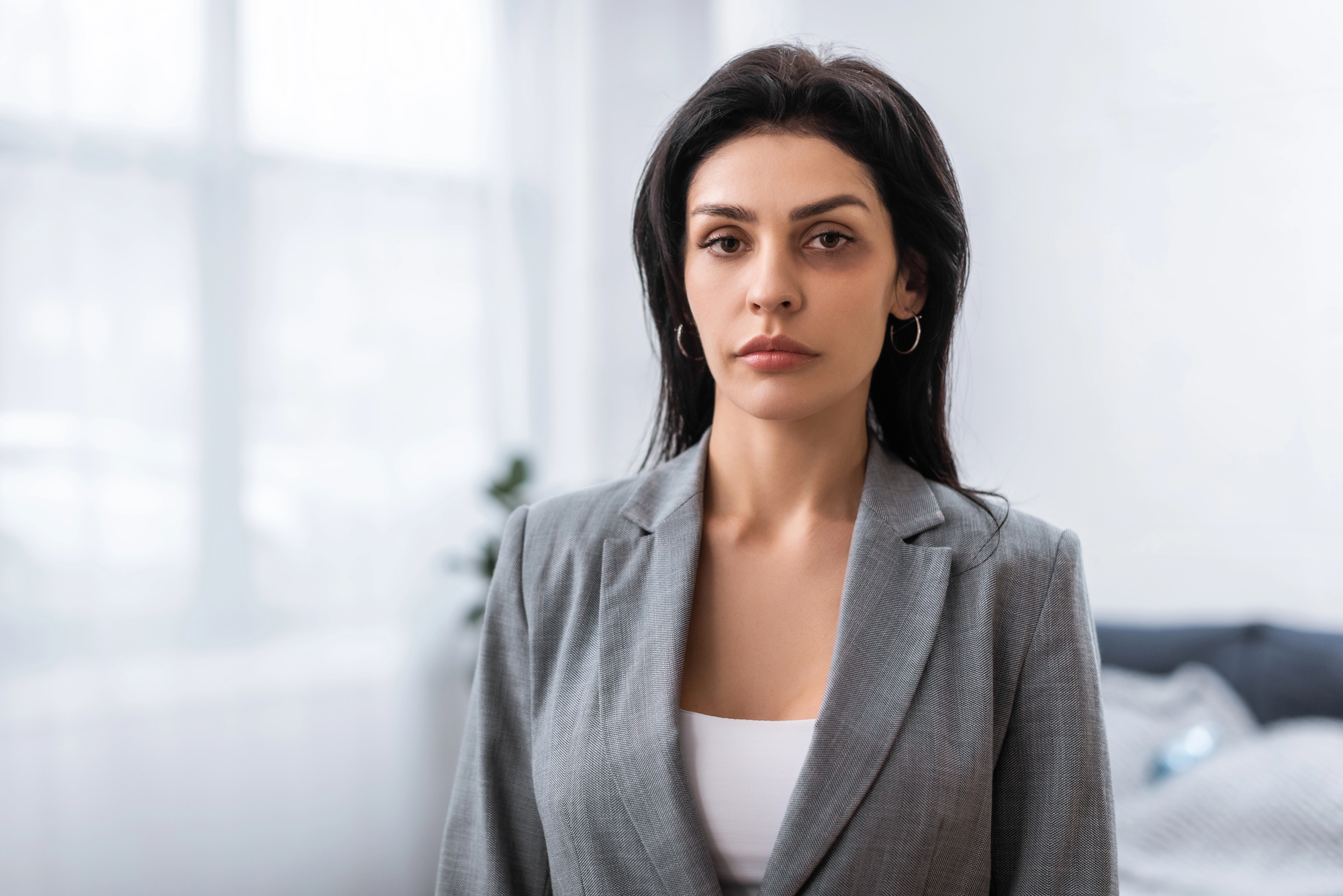 Woman with long dark hair and hoop earrings wearing a gray blazer stands in a softly lit room. She has a neutral expression and is facing the camera. The background features blurred curtains and hints of furniture.