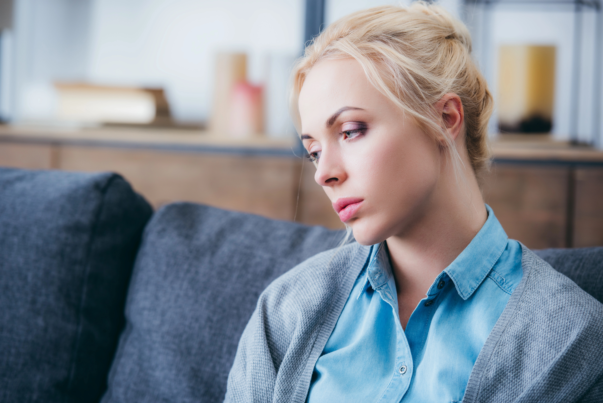 A woman with blonde hair is sitting on a gray couch, looking pensive. She is wearing a blue shirt and a gray cardigan. The background is softly blurred, with hints of furniture.