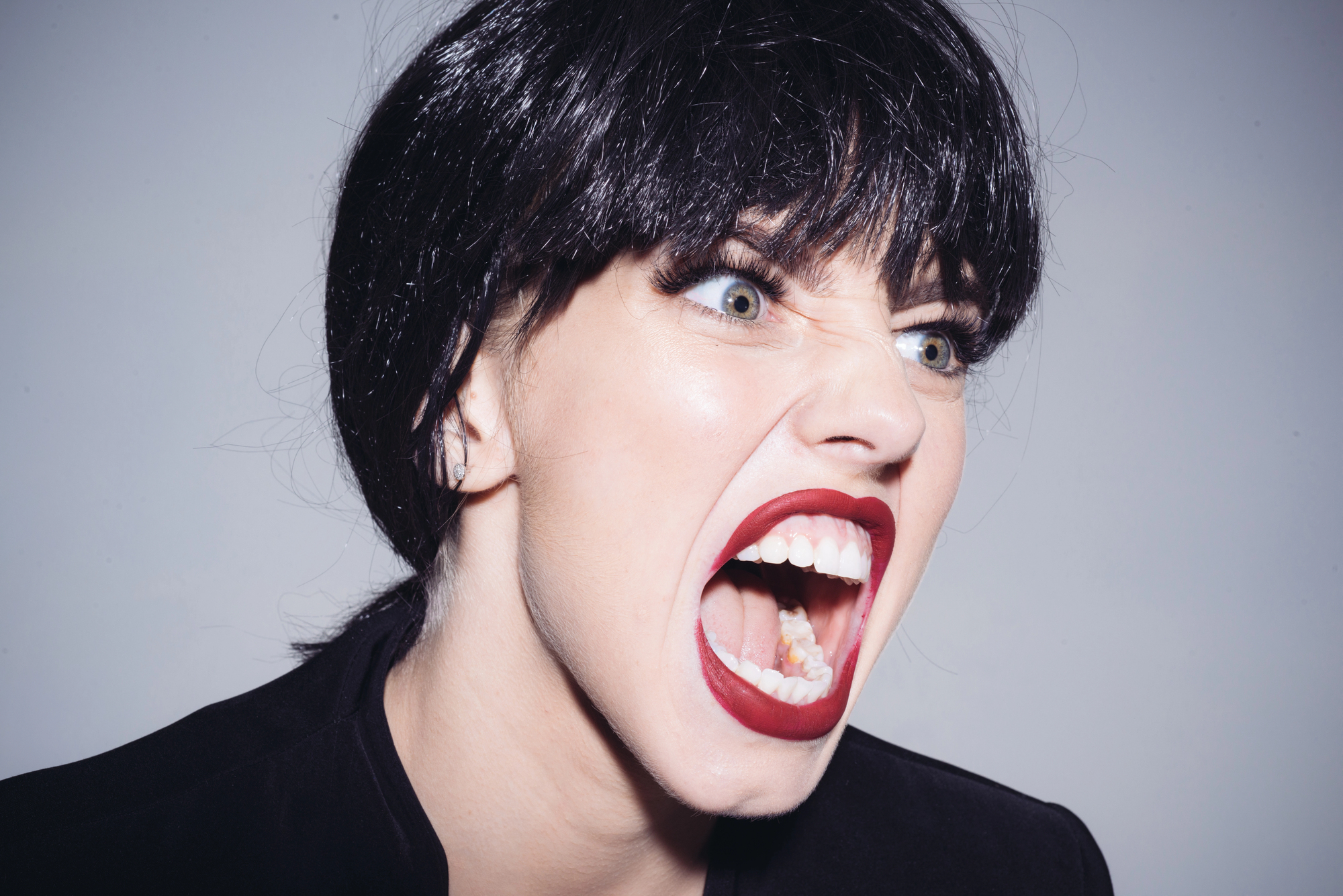 A person with short black hair and bold makeup is expressing anger or frustration. Their mouth is open wide, revealing teeth and a bright red lipstick. The background is plain, emphasizing their facial expression.