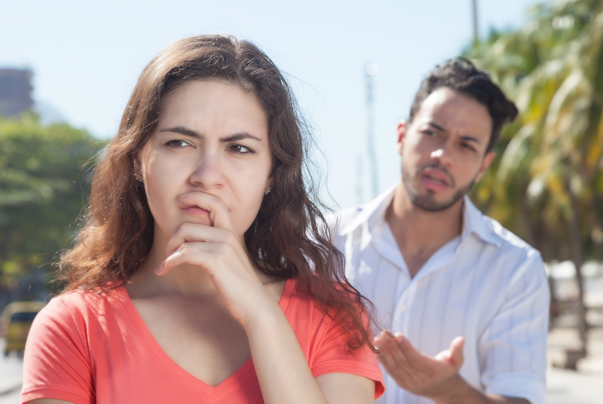 A woman in an orange shirt looks thoughtful and concerned, with her hand on her chin. Behind her, a man in a white shirt seems to be in a discussion, gesturing with one hand. They are outdoors on a sunny day.