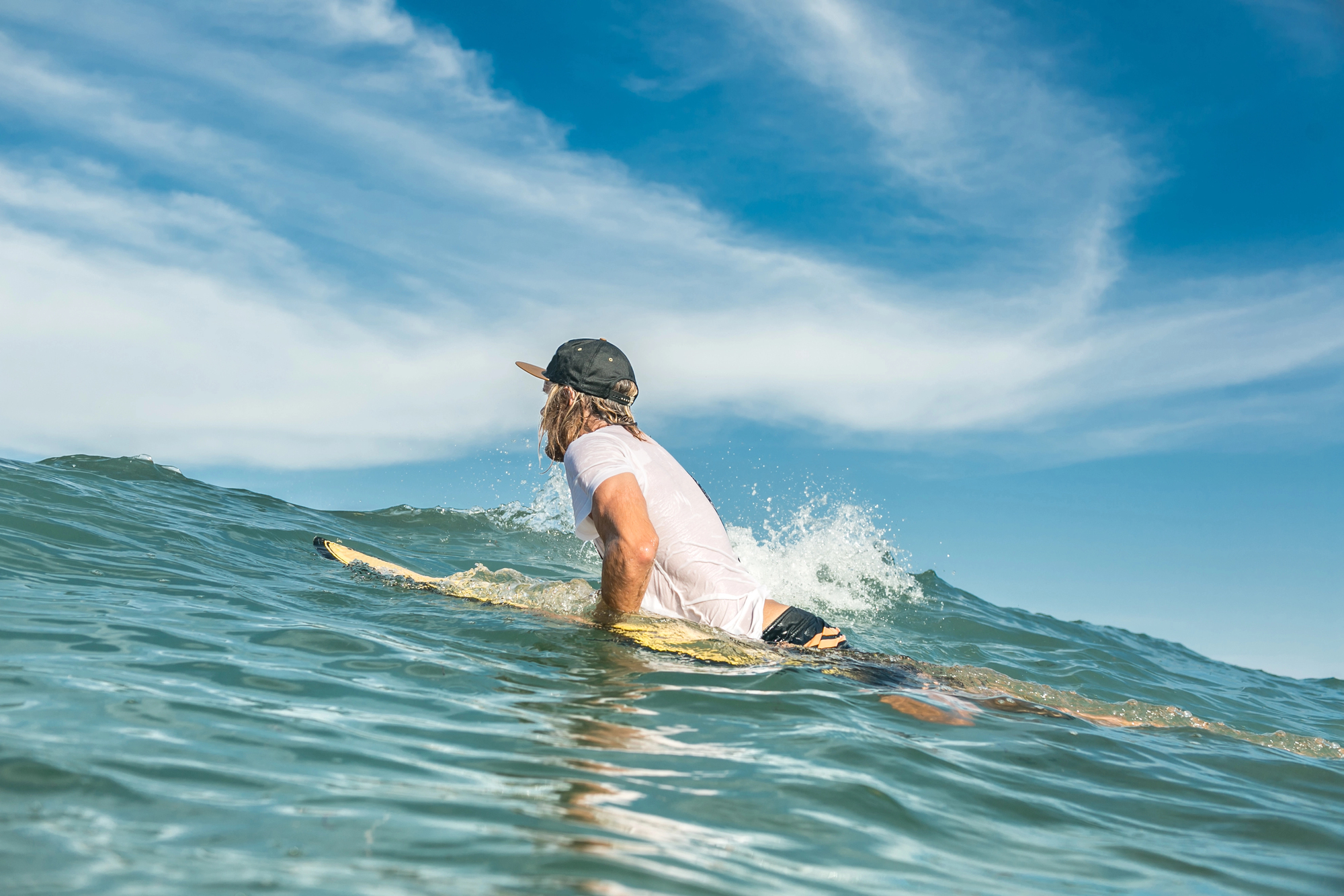 A person wearing a baseball cap and white shirt paddles on a surfboard through blue ocean waves under a clear sky with scattered clouds.