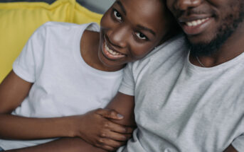 A couple sits closely on a yellow couch, both wearing white shirts. The woman leans her head on the man's shoulder, smiling warmly, while holding his arm. Their relaxed and happy demeanor creates a cozy and intimate atmosphere.