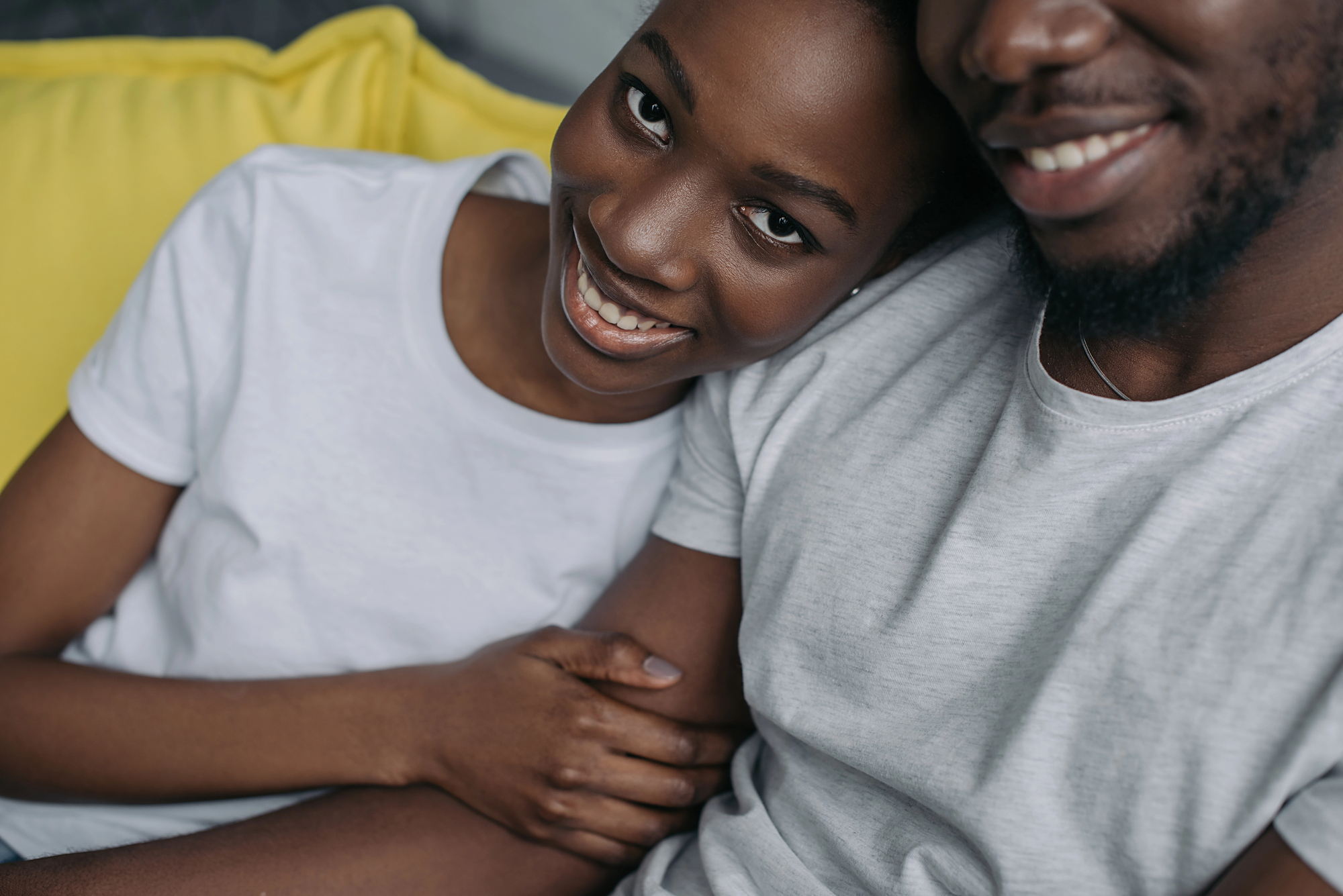 A couple sits closely on a yellow couch, both wearing white shirts. The woman leans her head on the man's shoulder, smiling warmly, while holding his arm. Their relaxed and happy demeanor creates a cozy and intimate atmosphere.