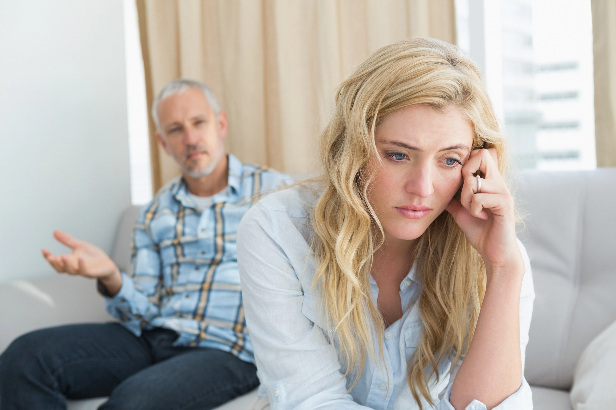 A woman with long blonde hair looks upset, resting her head on her hand, while sitting on a couch. In the background, a man with short gray hair and a checkered shirt gestures with one hand, seemingly talking to her.