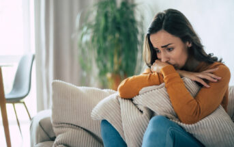 A woman sitting on a couch appears deep in thought, leaning on a pillow with a worried expression. She is dressed in a long-sleeve orange top and jeans. In the background, there's a blurred view of a plant and a table.