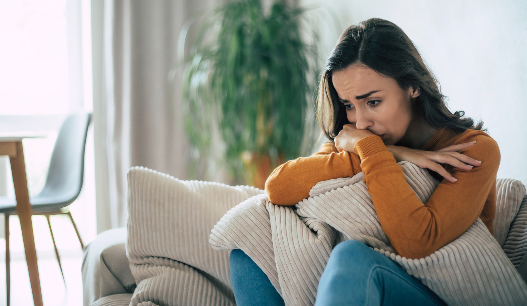 A woman sitting on a couch appears deep in thought, leaning on a pillow with a worried expression. She is dressed in a long-sleeve orange top and jeans. In the background, there's a blurred view of a plant and a table.