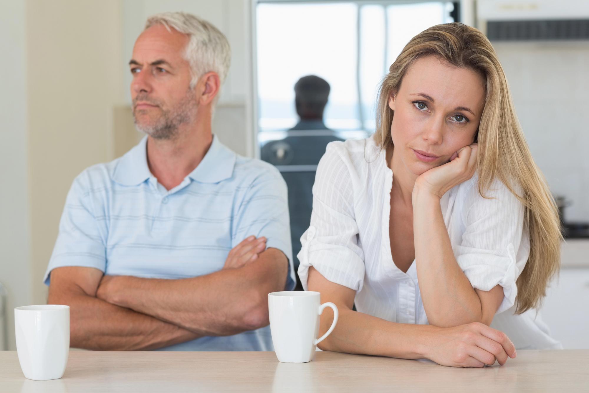 A man and a woman sit at a table with crossed arms. The woman, leaning on her hand, looks forward with a tired expression, while the man looks away. Two white mugs are on the table. The setting appears to be a kitchen.