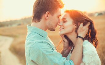 A couple stands in a warm embrace in a sunlit field. The man gently touches the woman's hair as they close their eyes and lean in close. The setting sun casts a golden glow, creating a romantic and serene atmosphere.