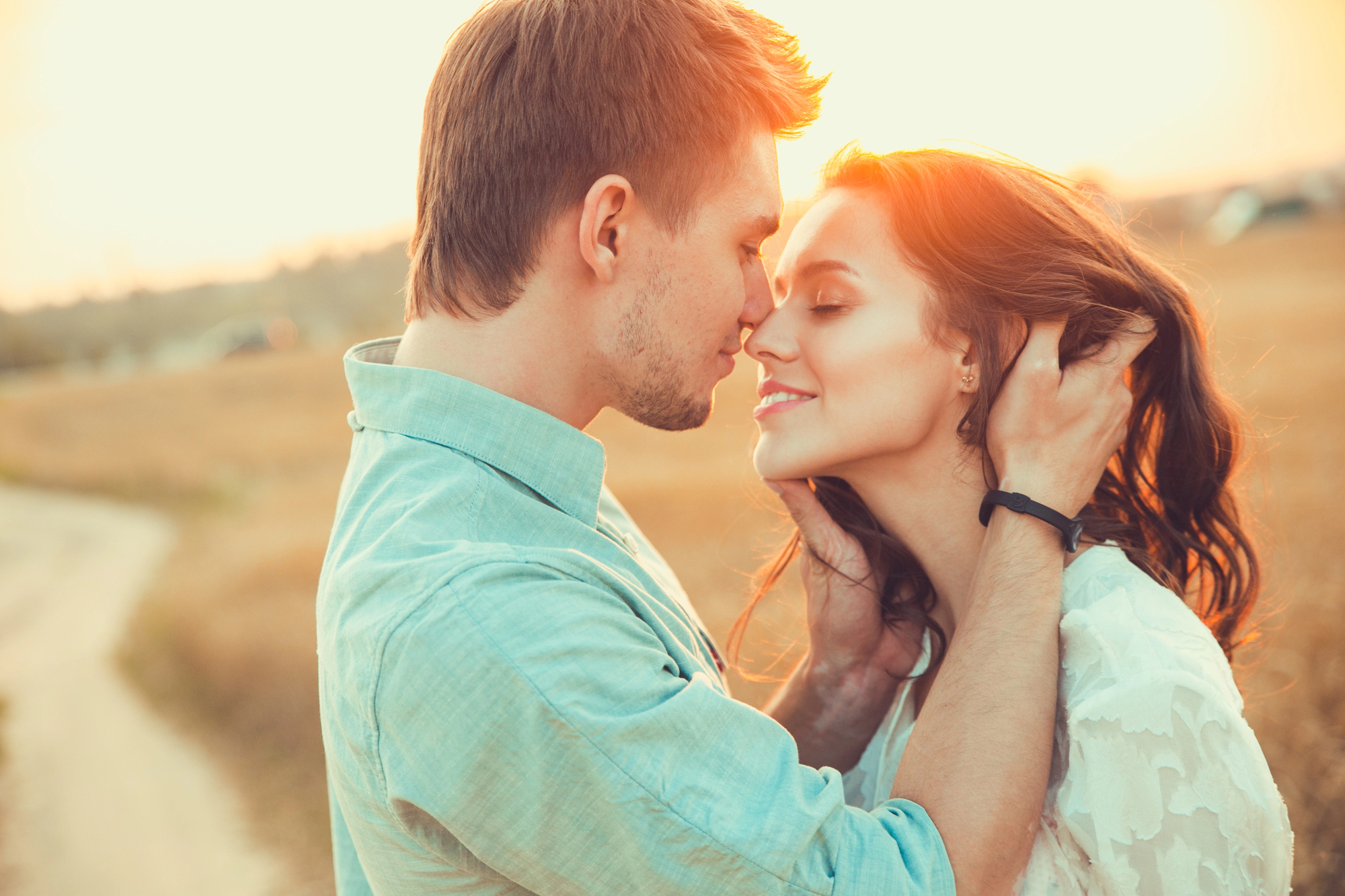 A couple stands in a warm embrace in a sunlit field. The man gently touches the woman's hair as they close their eyes and lean in close. The setting sun casts a golden glow, creating a romantic and serene atmosphere.