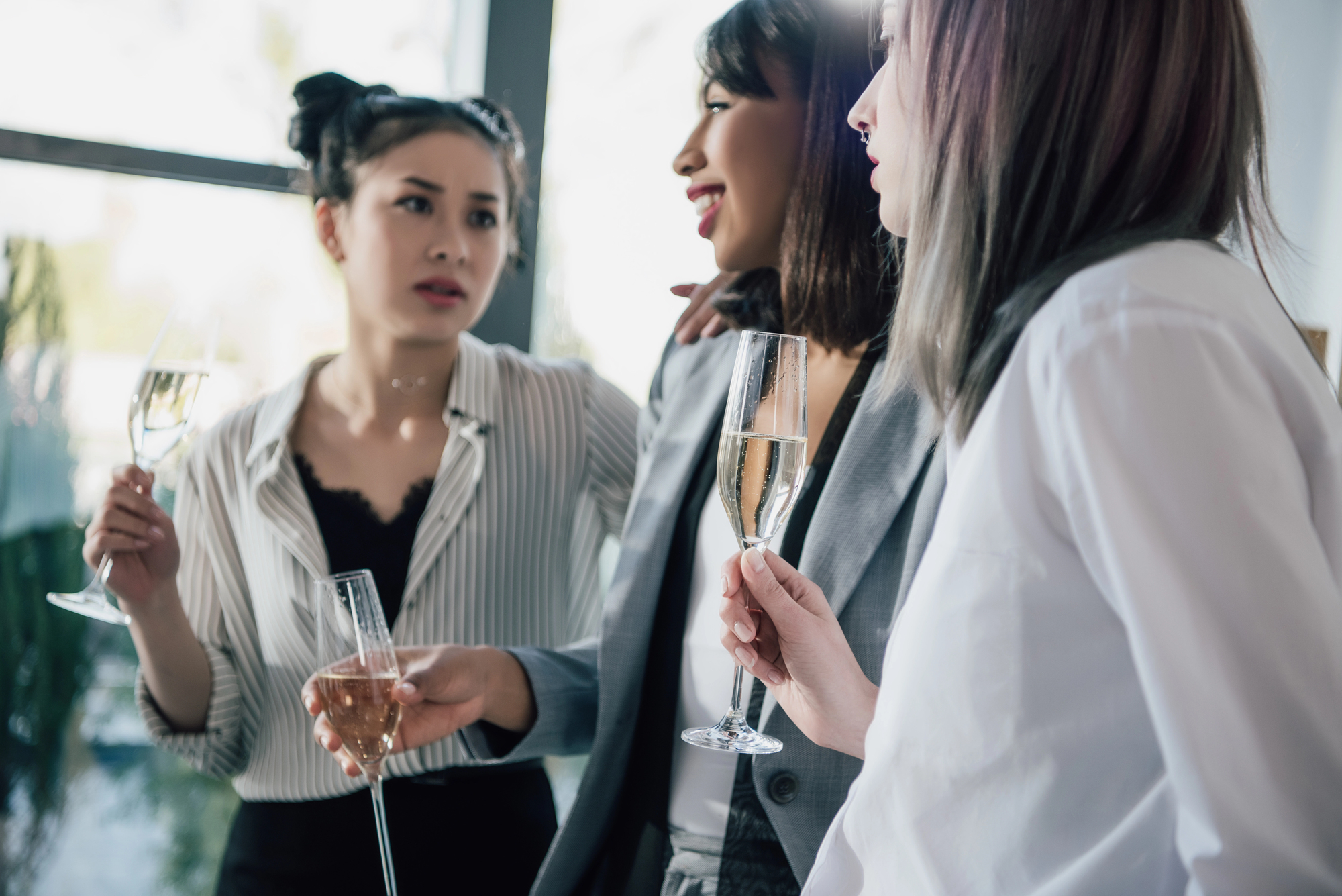 Three women are socializing indoors, each holding a glass of champagne. They appear engaged in conversation, with natural light streaming in from a window behind them. One woman wears a striped shirt, another a gray blazer, and the third a white shirt.