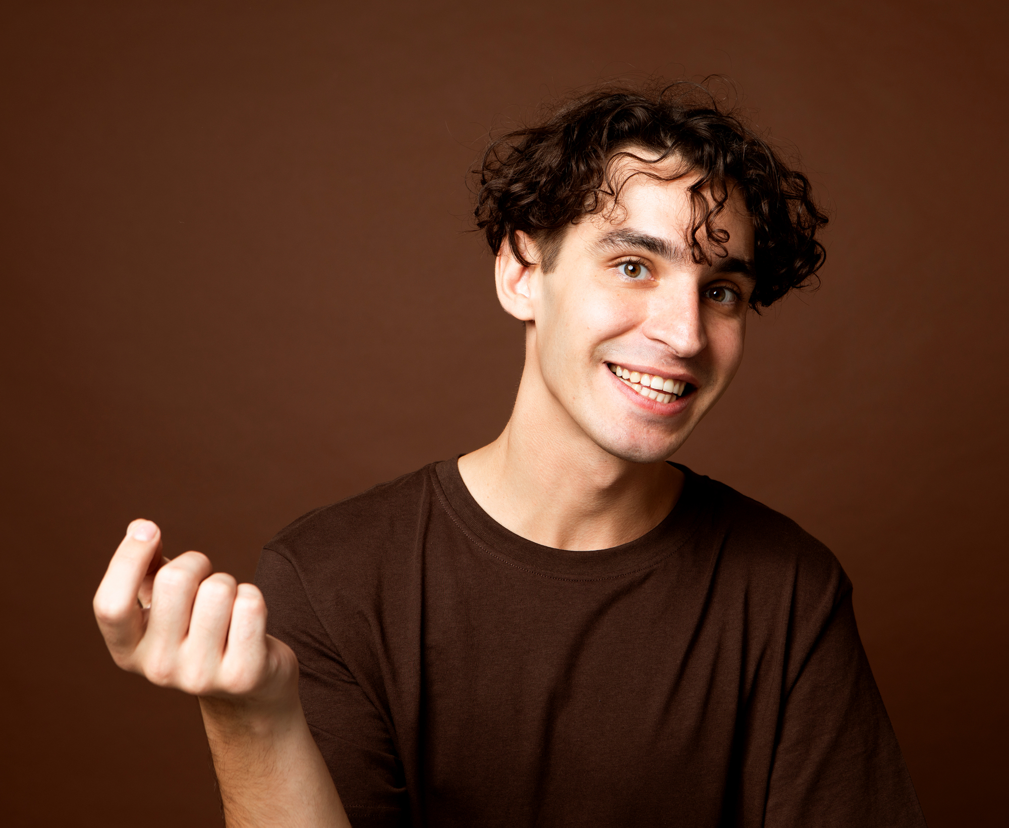 A person with curly hair is smiling broadly against a brown background, wearing a brown shirt. One hand is raised in a casual gesture.