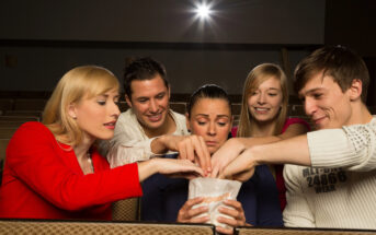 Five people sitting in a movie theater share popcorn. They are reaching into a single popcorn bag, smiling and engaged with each other in a friendly atmosphere. The theater seats surround them, and the lighting is dim.