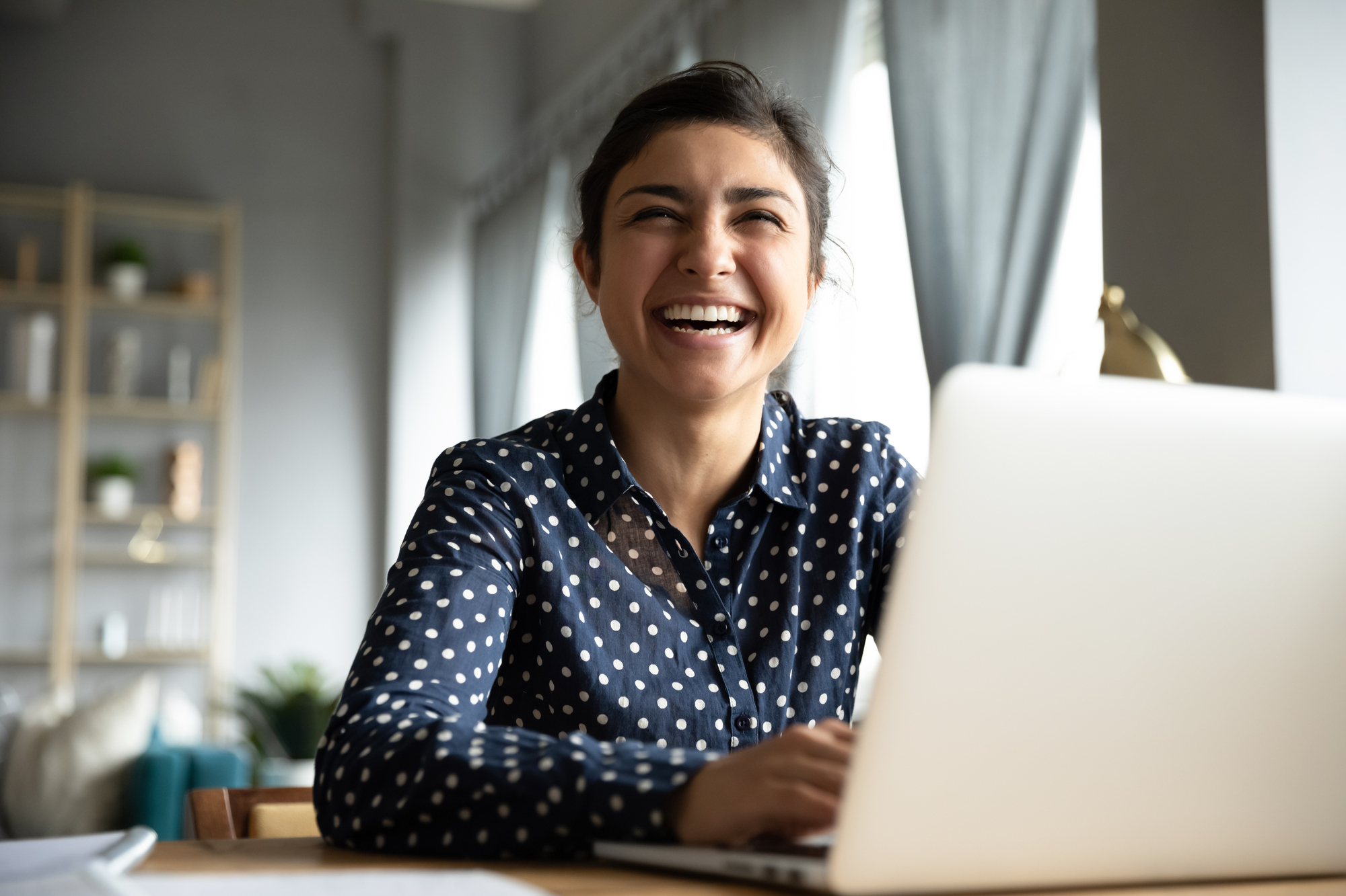 A woman in a polka dot blouse is sitting at a table, smiling broadly while using a laptop. Behind her are shelves and large windows with curtains, creating a bright and cozy atmosphere.