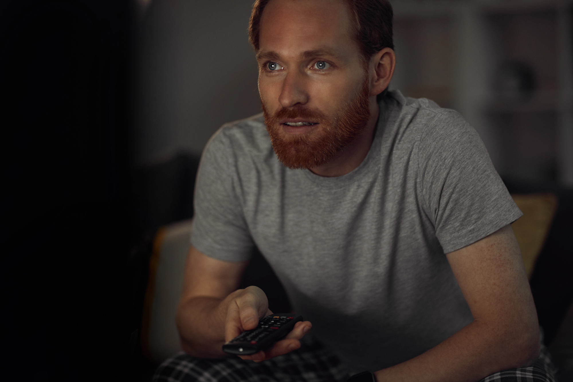 A man with a beard and casually dressed in a gray t-shirt and checkered pants sits on a couch holding a remote control, focused on a TV screen in a dimly lit room.