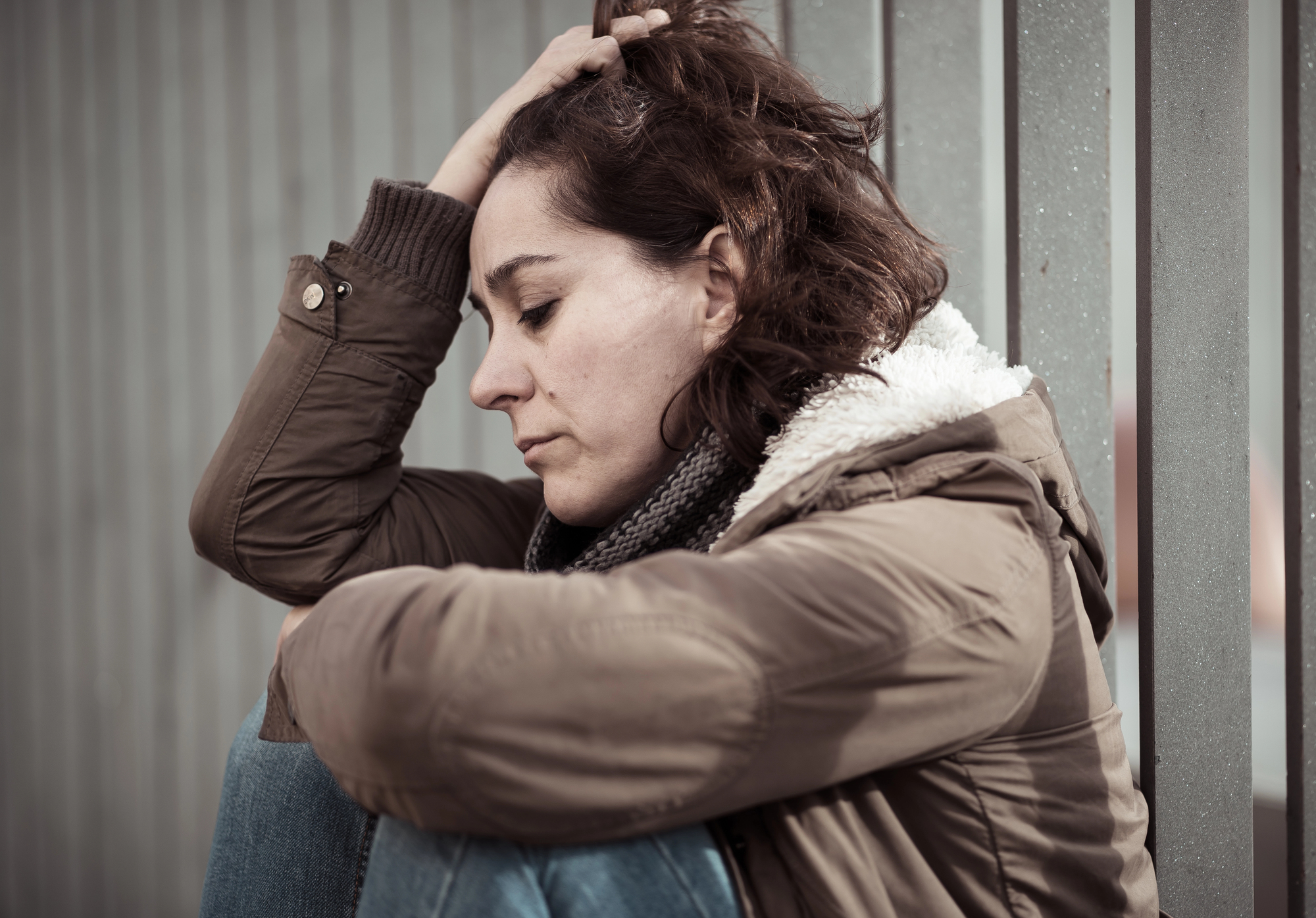 A woman sitting against a metal fence, looking contemplative. She is wearing a brown coat, a scarf, and jeans. Her hand rests on her head, and her expression seems thoughtful and reflective.