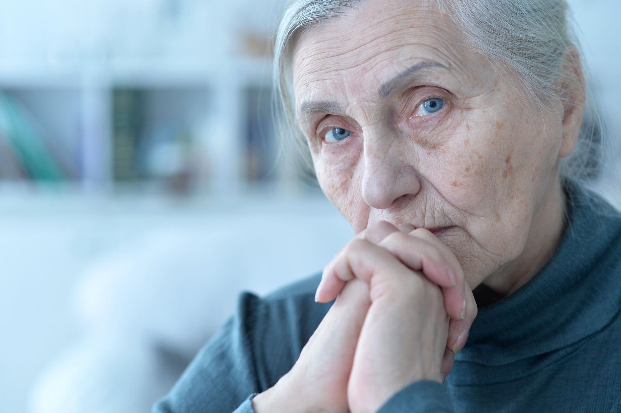 Elderly woman with gray hair and a thoughtful expression sits with her hands clasped near her face, looking directly at the camera. She wears a gray turtleneck sweater, and the background is softly blurred, with hints of books on shelves.