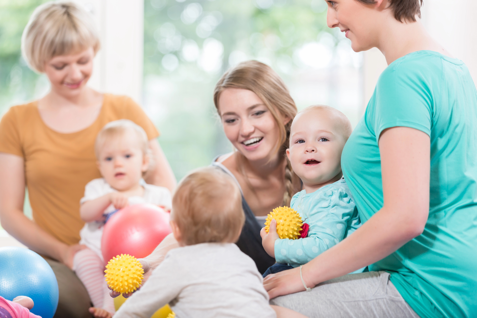 Three women and three toddlers are sitting together in a bright room. The women are smiling, and the toddlers are holding colorful balls. One toddler has a yellow spiky ball while another toddler plays nearby. The atmosphere is cheerful and playful.