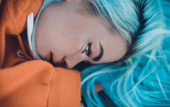 Close-up of a person with vibrant blue hair lying down, wearing an orange hoodie. They gaze thoughtfully into the distance, creating a striking contrast between their hair and clothing.