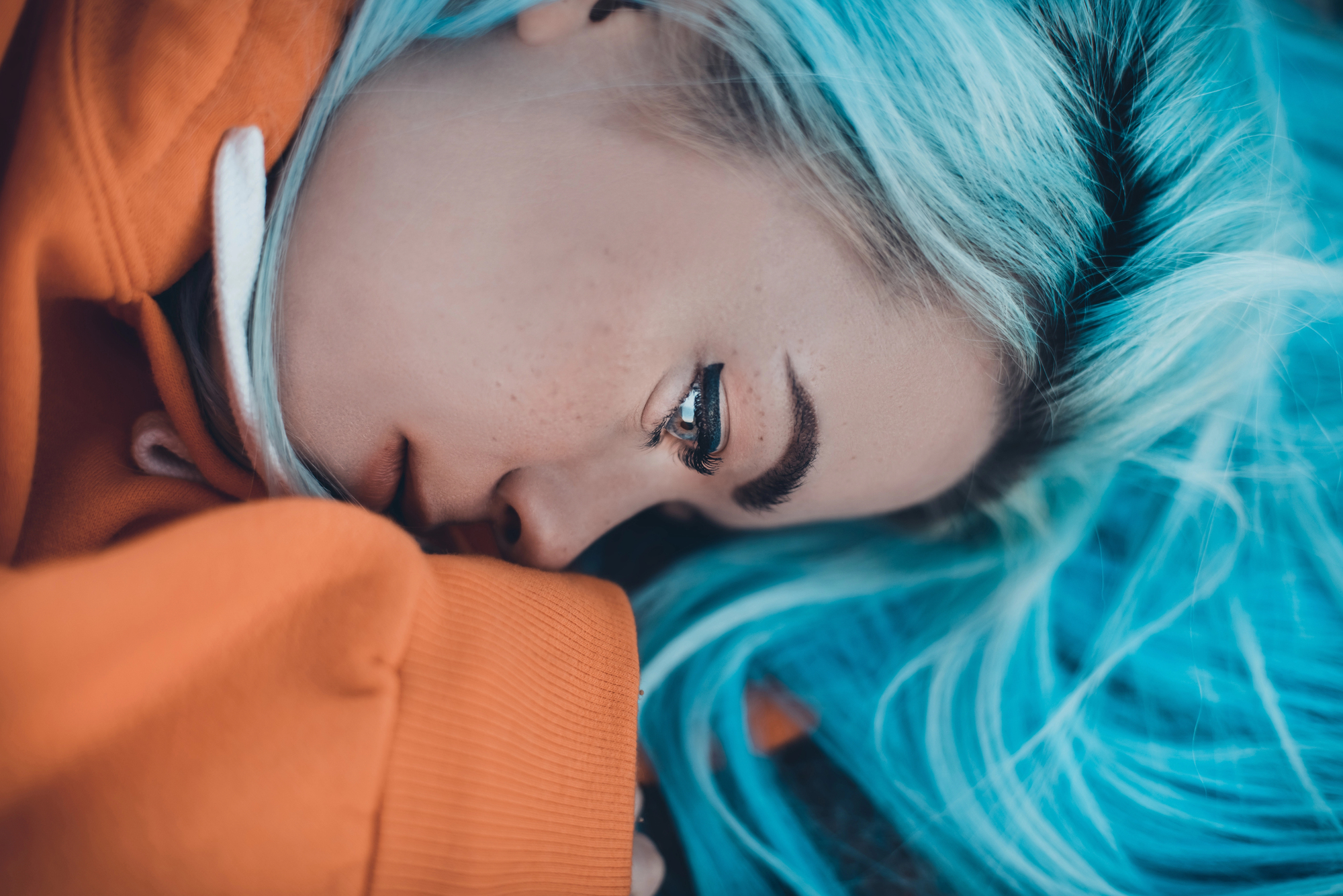 Close-up of a person with vibrant blue hair lying down, wearing an orange hoodie. They gaze thoughtfully into the distance, creating a striking contrast between their hair and clothing.