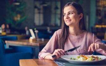 A woman with long brown hair is sitting at a restaurant table, smiling and holding a fork and knife. She is about to eat a meal on a white plate. The background shows a cozy, blurred restaurant setting with blue chairs.