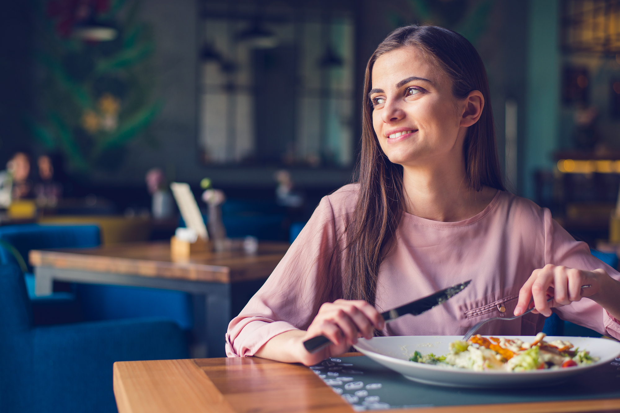 A woman with long brown hair is sitting at a restaurant table, smiling and holding a fork and knife. She is about to eat a meal on a white plate. The background shows a cozy, blurred restaurant setting with blue chairs.