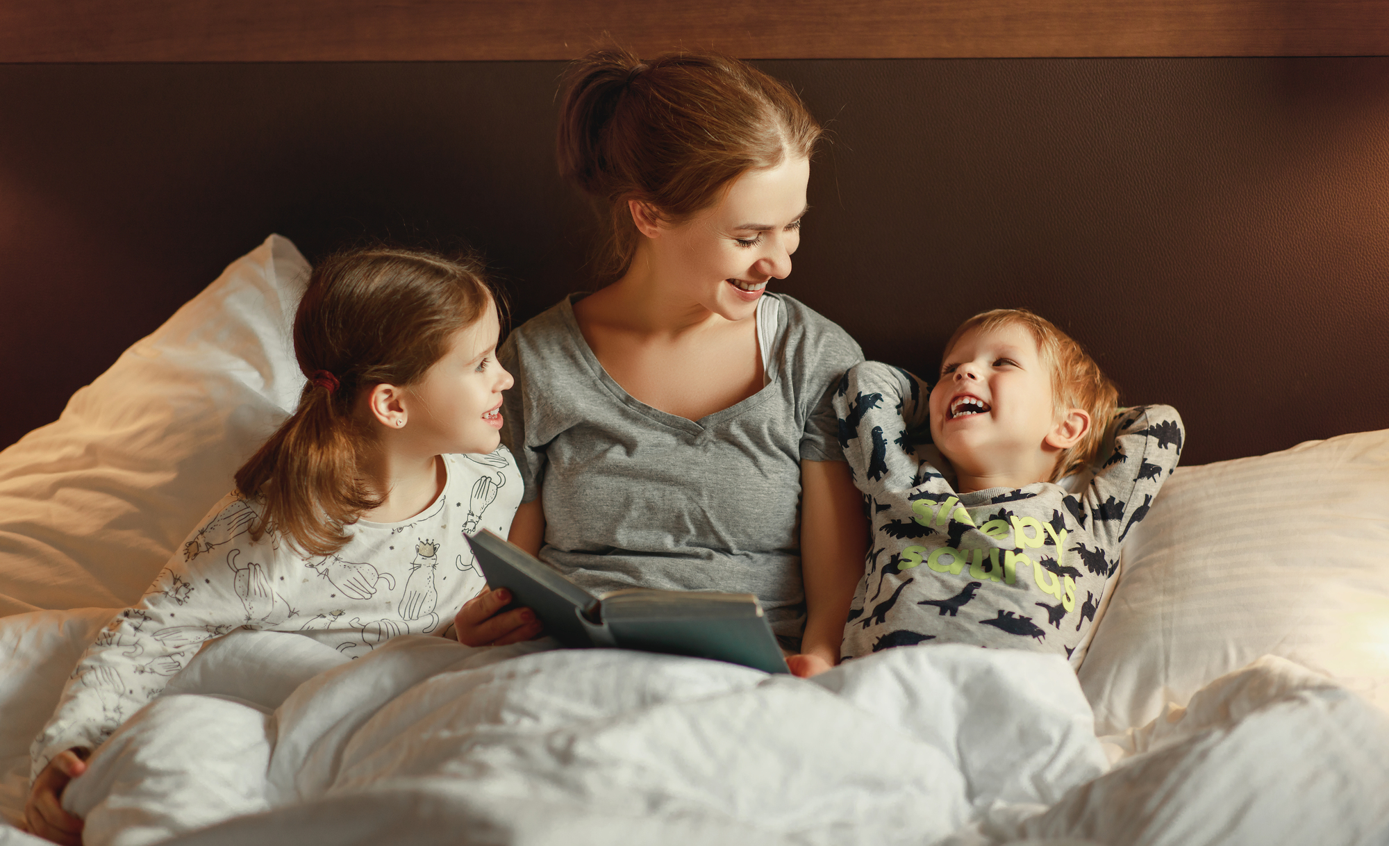 A smiling woman sits in bed with two young children. One child is leaning towards her while the other is lying back with hands behind their head. They all appear happy and are surrounded by pillows and soft lighting.