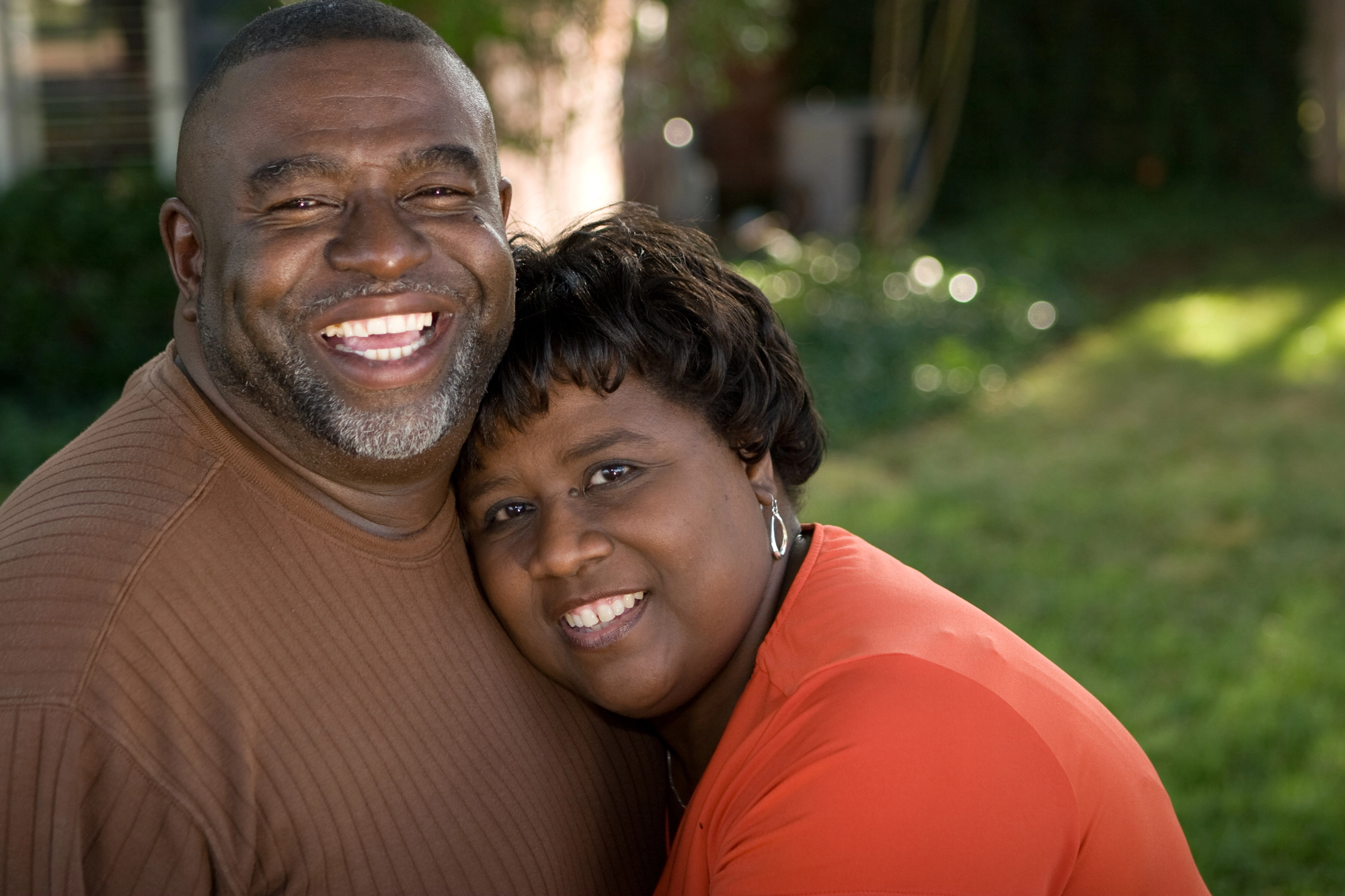 A smiling couple embraces outdoors. The man, wearing a brown sweater, has a gray beard. The woman, in an orange top, leans her head on his shoulder. They appear happy, with a blurred green background behind them.