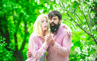 A couple stands close together in a lush green park, both blowing on small white dandelion seeds. The woman with blonde hair and a man with a beard wear light pink shirts. A flowering tree accentuates the serene spring setting.