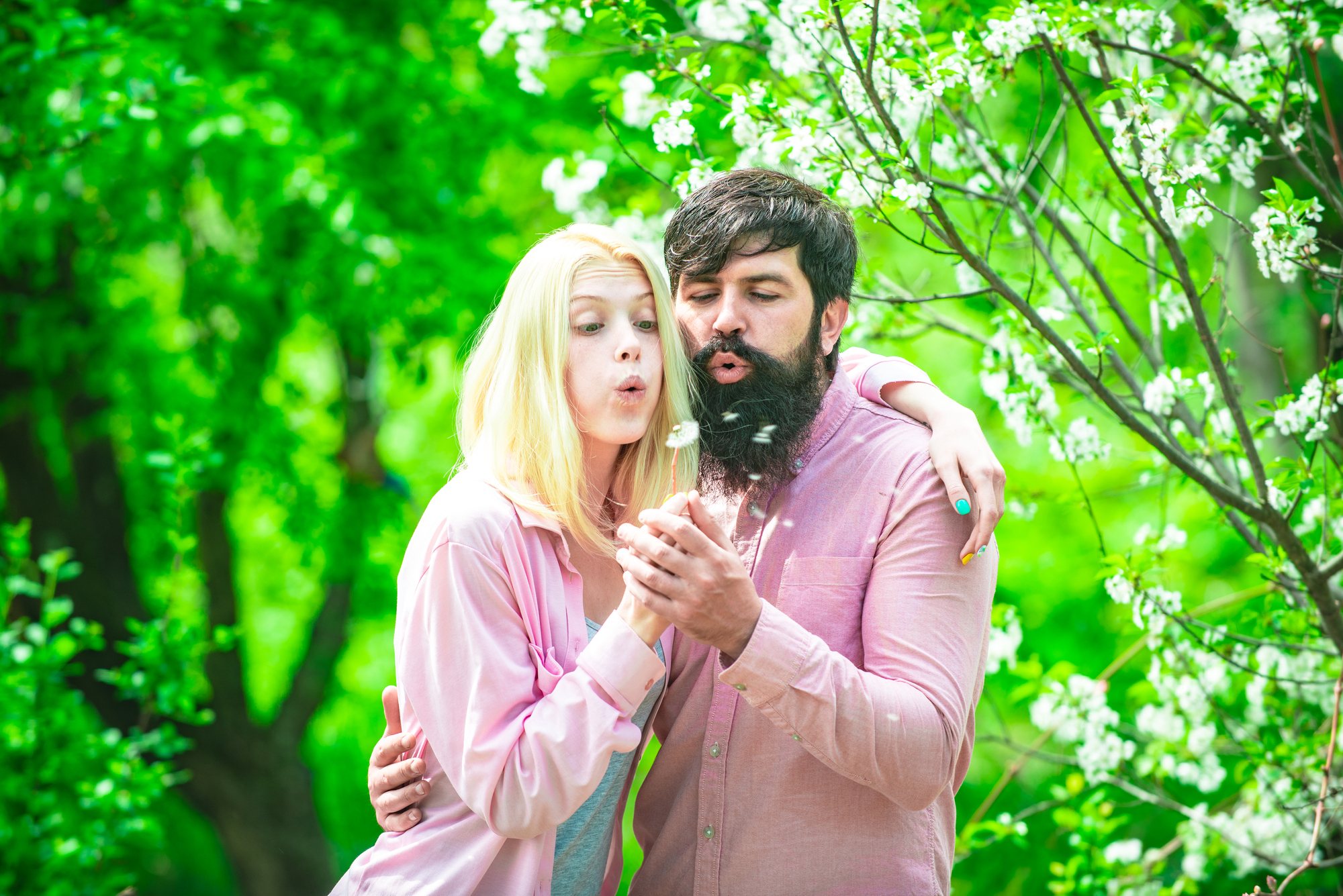 A couple stands close together in a lush green park, both blowing on small white dandelion seeds. The woman with blonde hair and a man with a beard wear light pink shirts. A flowering tree accentuates the serene spring setting.