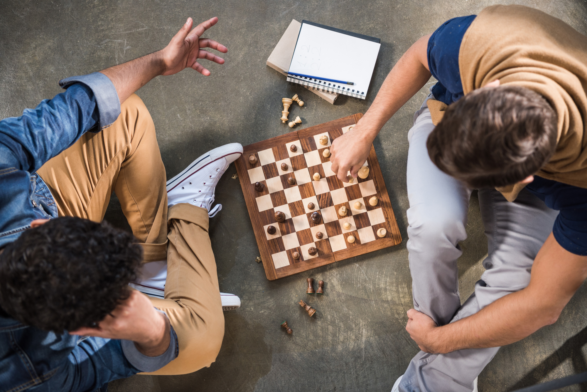 Two people sit on the floor playing chess. One has a notepad nearby. They are casually dressed, with one player wearing sneakers and the other with a sweater draped over their shoulders. Some chess pieces are scattered around the board.