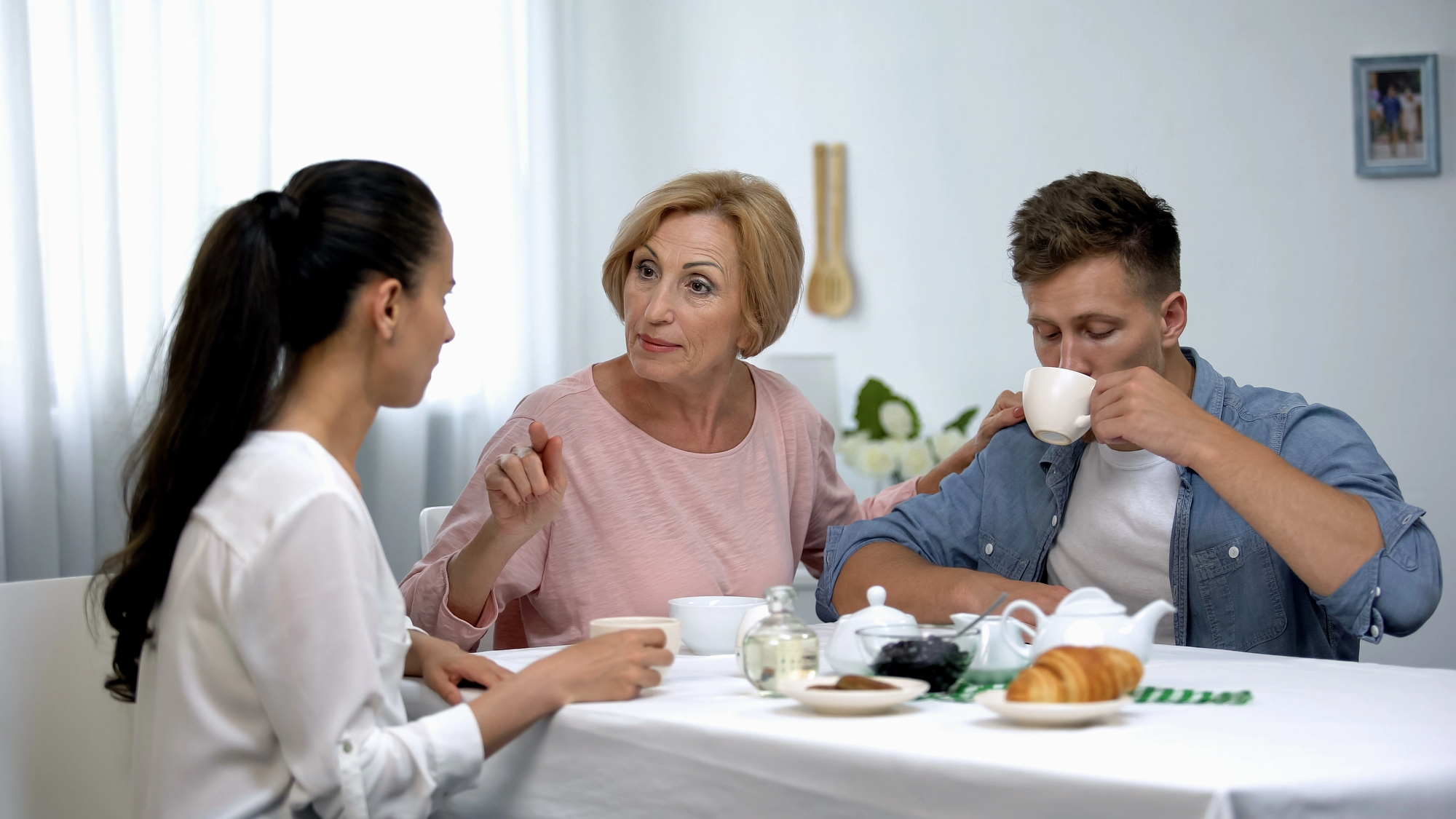 Three people sitting at a table with coffee cups: a woman with dark hair, an older woman speaking and gesturing, and a man sipping from a cup. A teapot, jam, and croissants are on the table in a well-lit room.