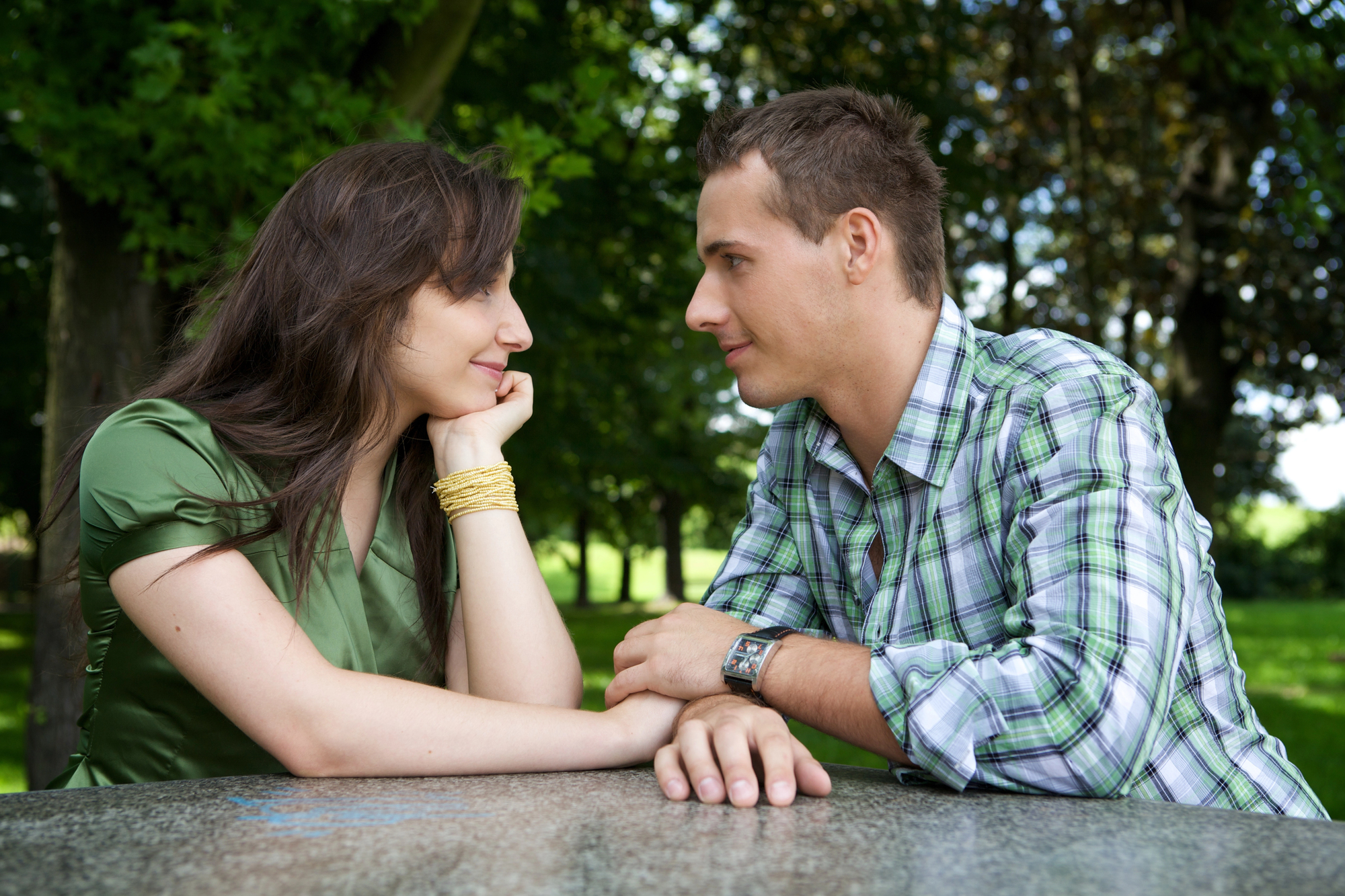 A man and woman sit across from each other at a stone table in a park, smiling and gazing into each other's eyes. The woman rests her chin on her hand, wearing a green blouse; the man wears a plaid shirt, and trees are in the background.